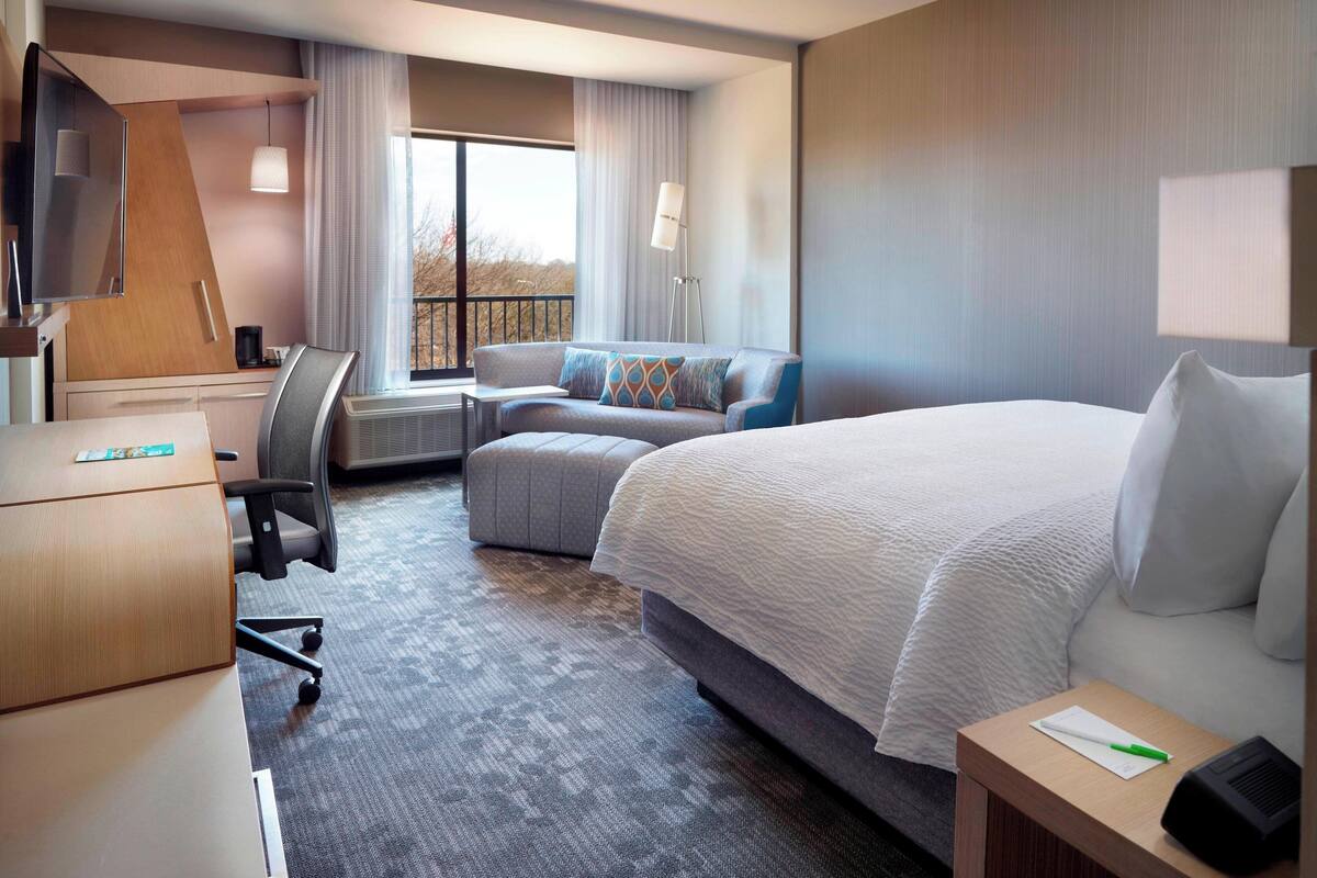 Quarto de hotel com uma cama grande, uma escrivaninha com computador, um sofá e uma janela com cortinas.
