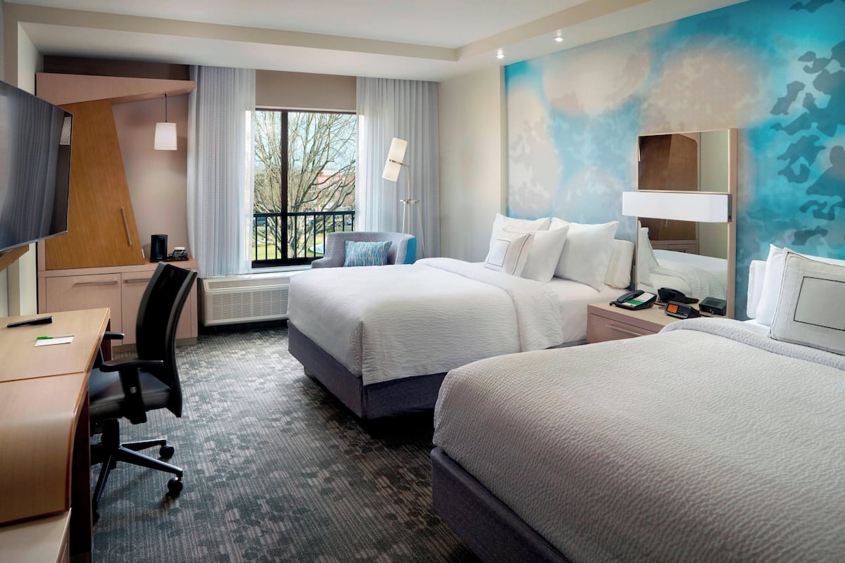 Quarto de hotel com duas camas, uma escrivaninha, uma cadeira, uma televisão e janela com vista para árvores.