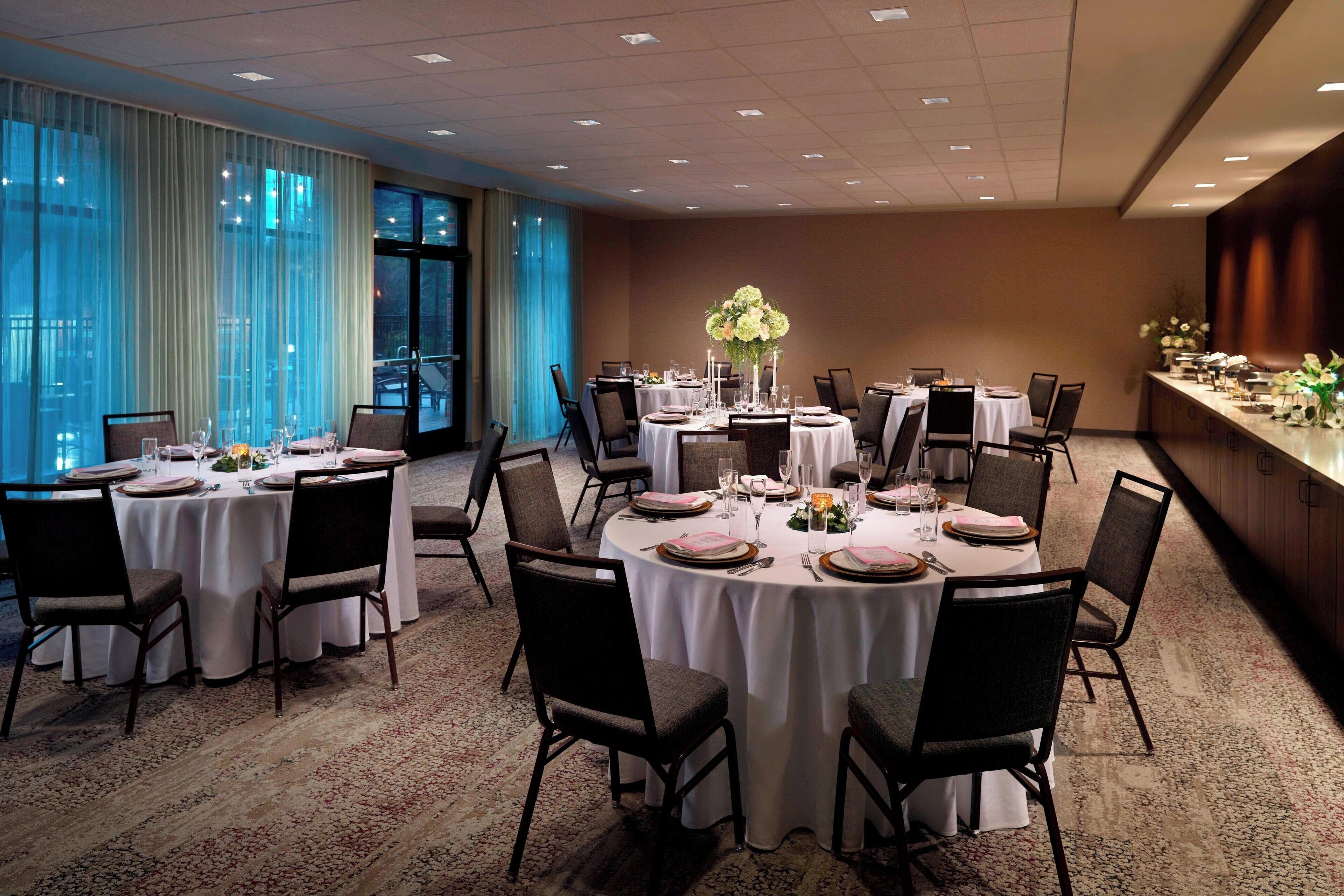 Un salón de banquetes con mesas redondas dispuestas para cenar, ventanas amplias con cortinas y vistas a la ciudad.