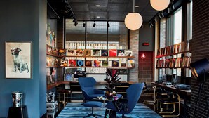 A modern interior with a long table, two blue chairs, a bookshelf filled with books, and a framed artwork on the wall. - MOXY Minneapolis Uptown (Minneapolis)