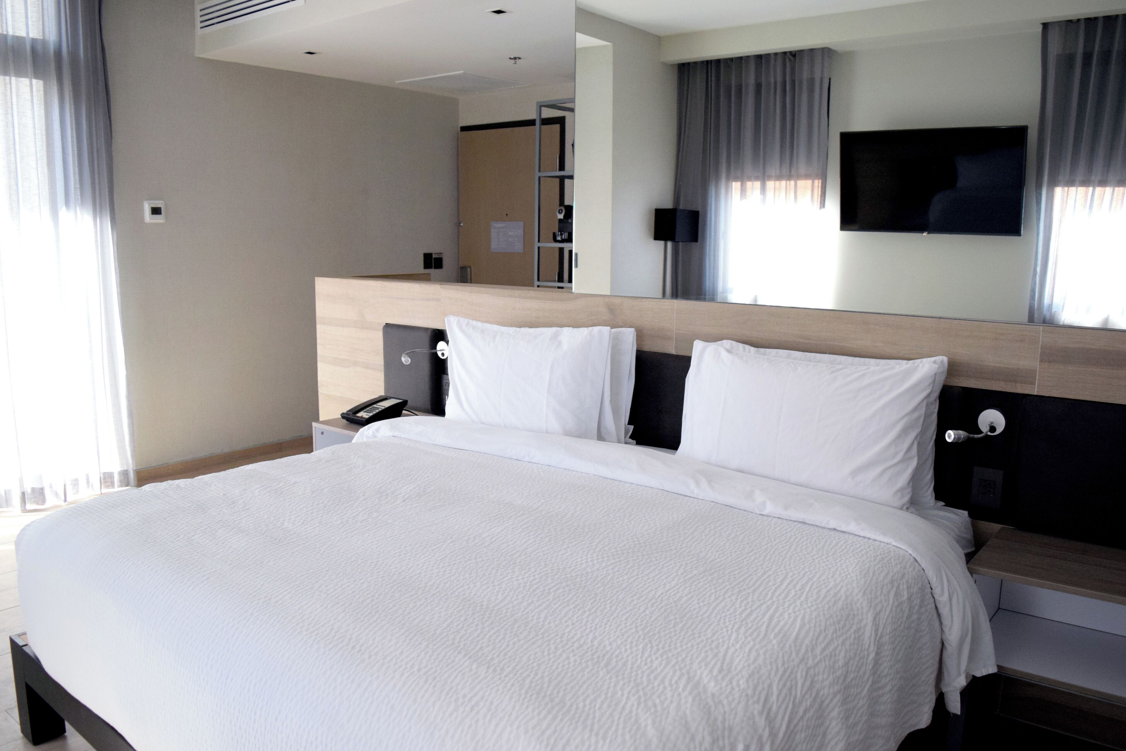 A hotel room with a large bed, a bedside table, a flat-screen TV mounted on the wall, and a window with curtains.