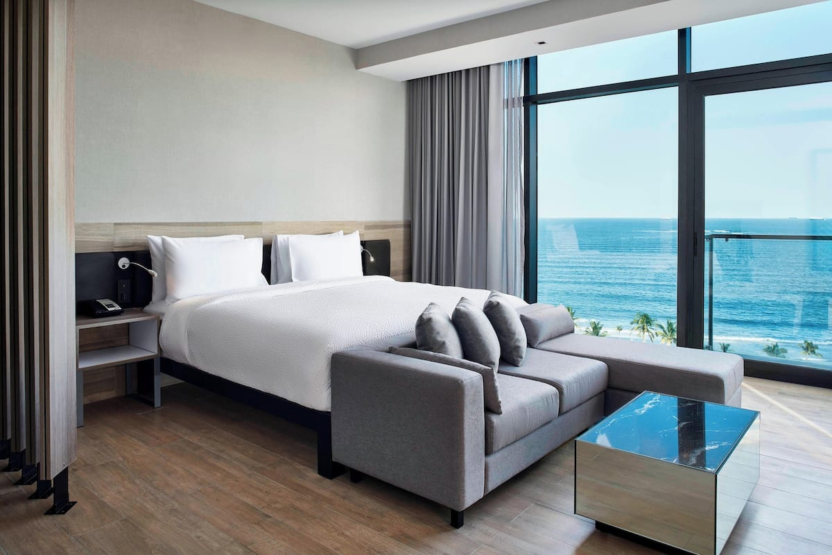 Una habitación de hotel moderna con una cama grande, un sofá, una mesa de centro de vidrio y vistas al océano.