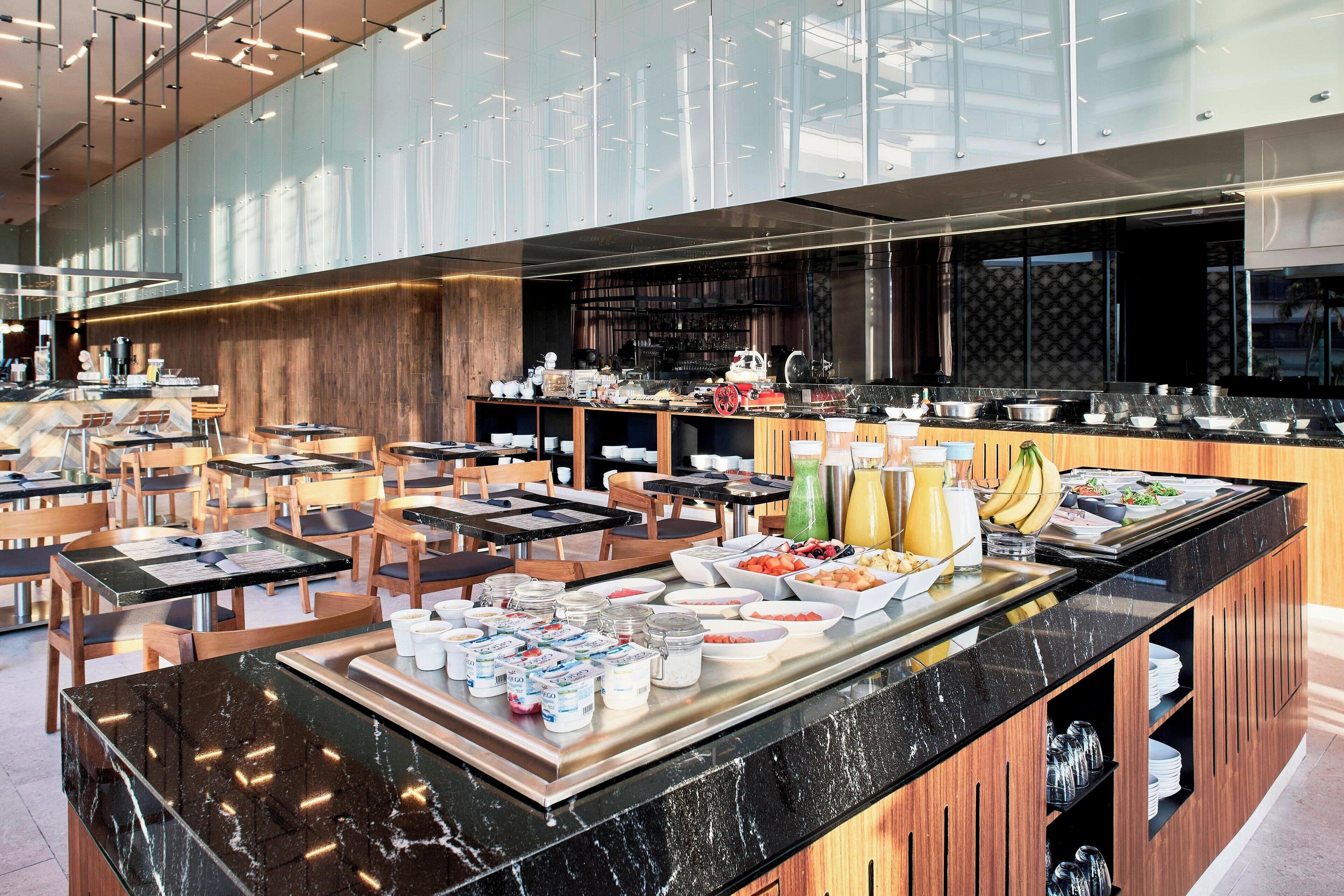 Et moderne hotelmorgenmadsområde med buffet, siddepladser og et synligt køkken i baggrunden.
