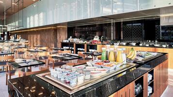 Et moderne hotelmorgenmadsområde med buffet, siddepladser og et synligt køkken i baggrunden.