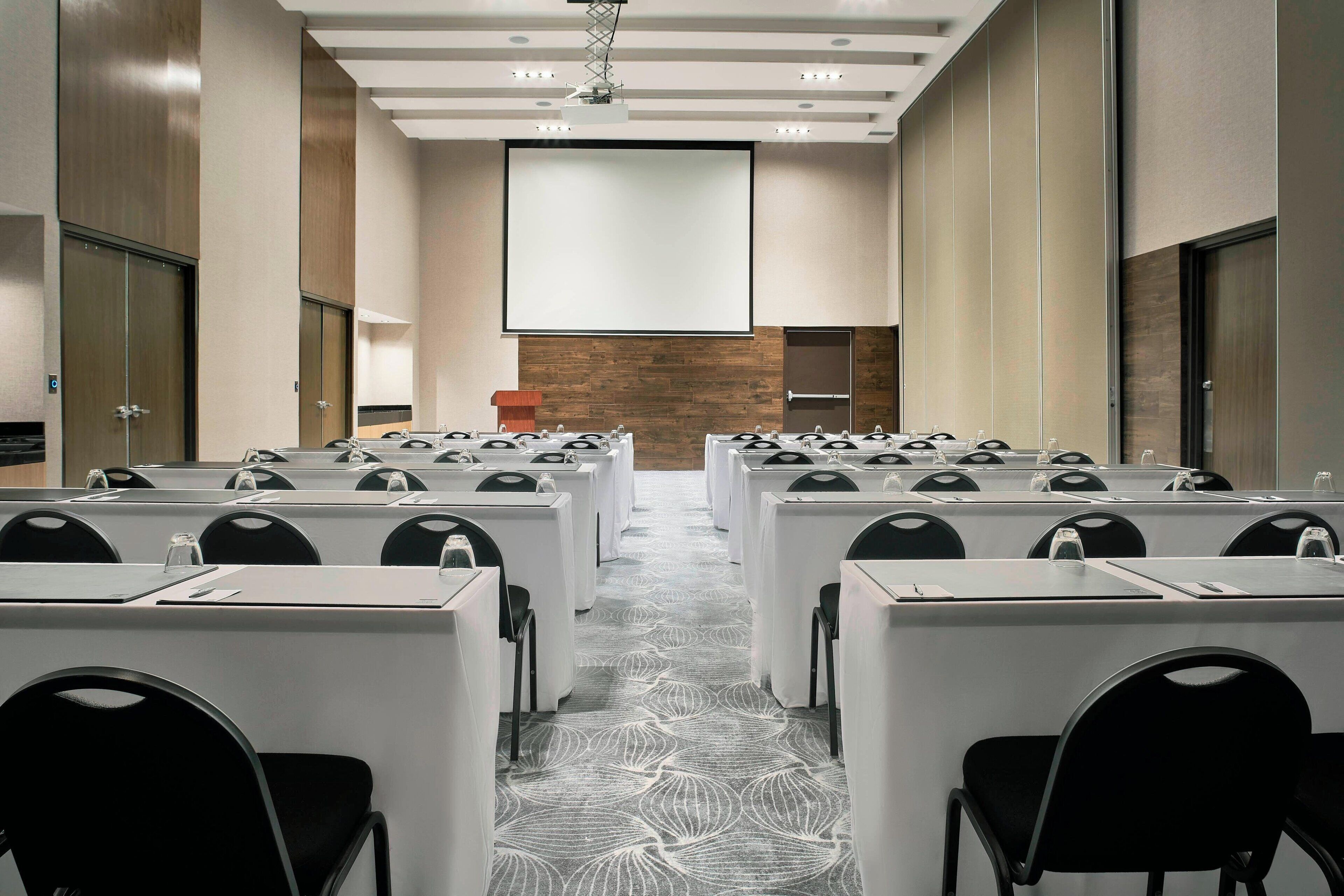A conference room with rows of tables and chairs, a large screen at the front, and a wooden accent wall.