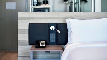 A hotel room with a bed, a nightstand with a phone and a small table, and a mirror reflecting a wardrobe and a shoe rack.