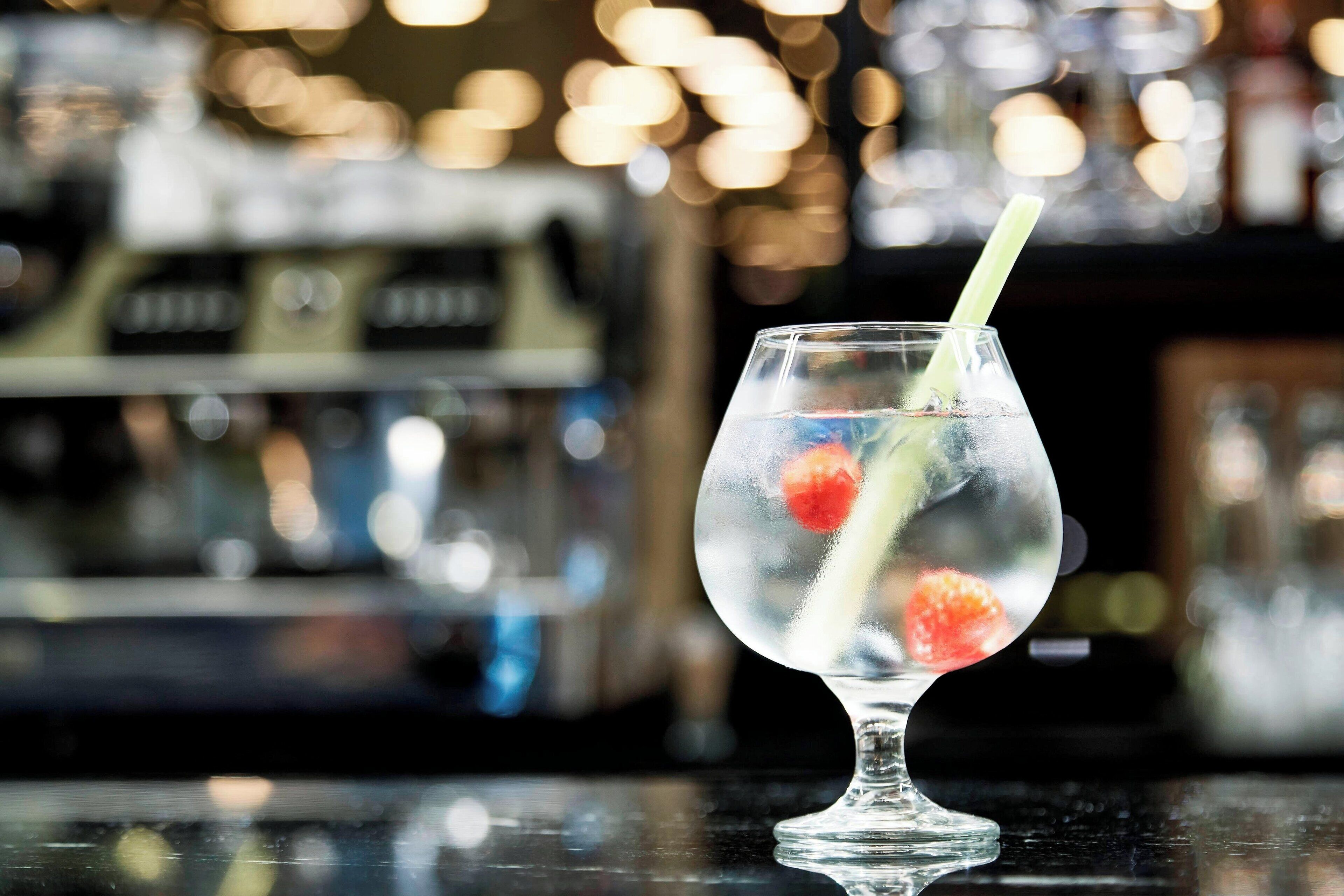 A clear glass with a straw, containing ice and a red fruit, on a bar counter with a blurred background.
