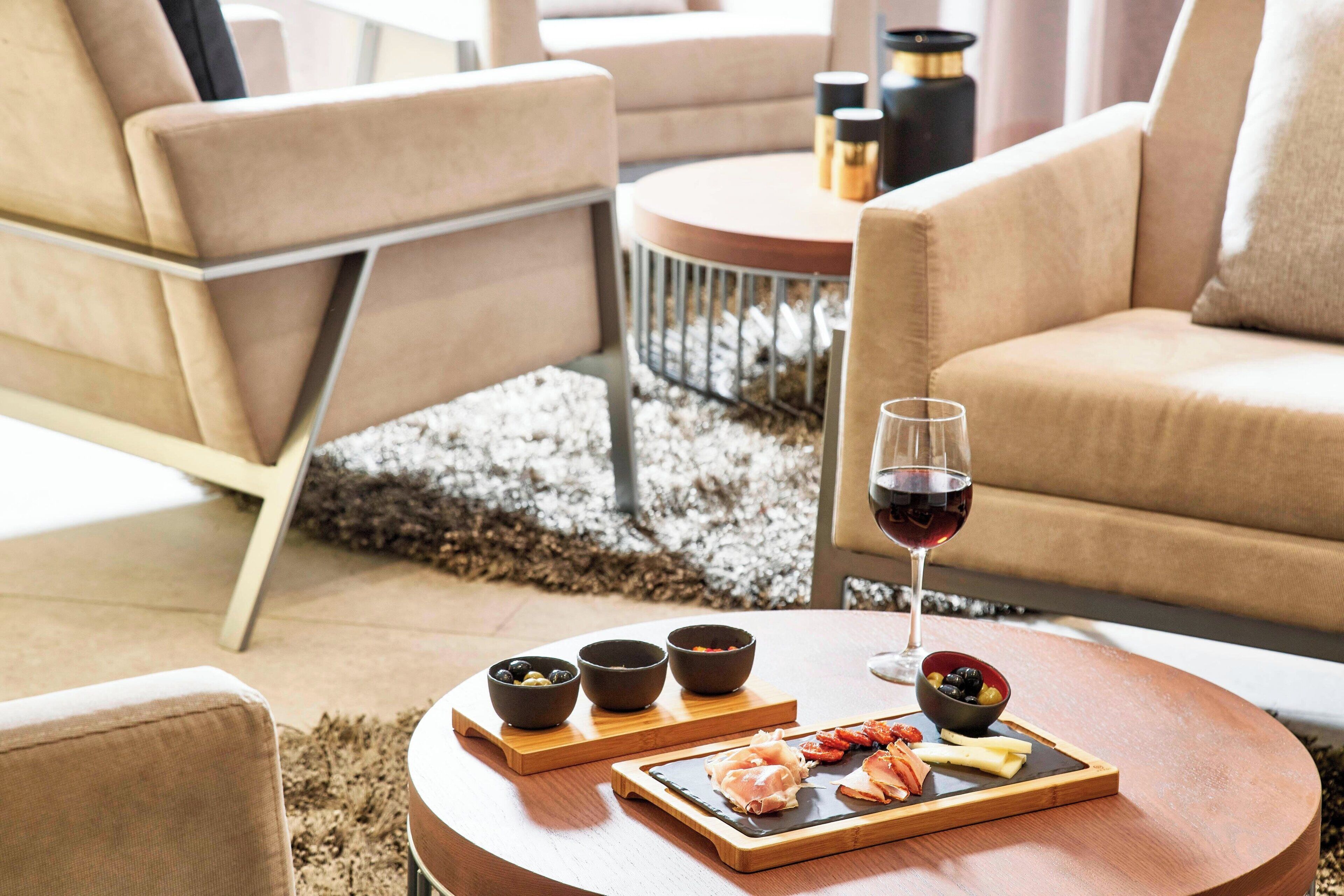 A modern living room with a round coffee table, beige sofas, and a small dining area with a wooden board displaying food and drinks.
