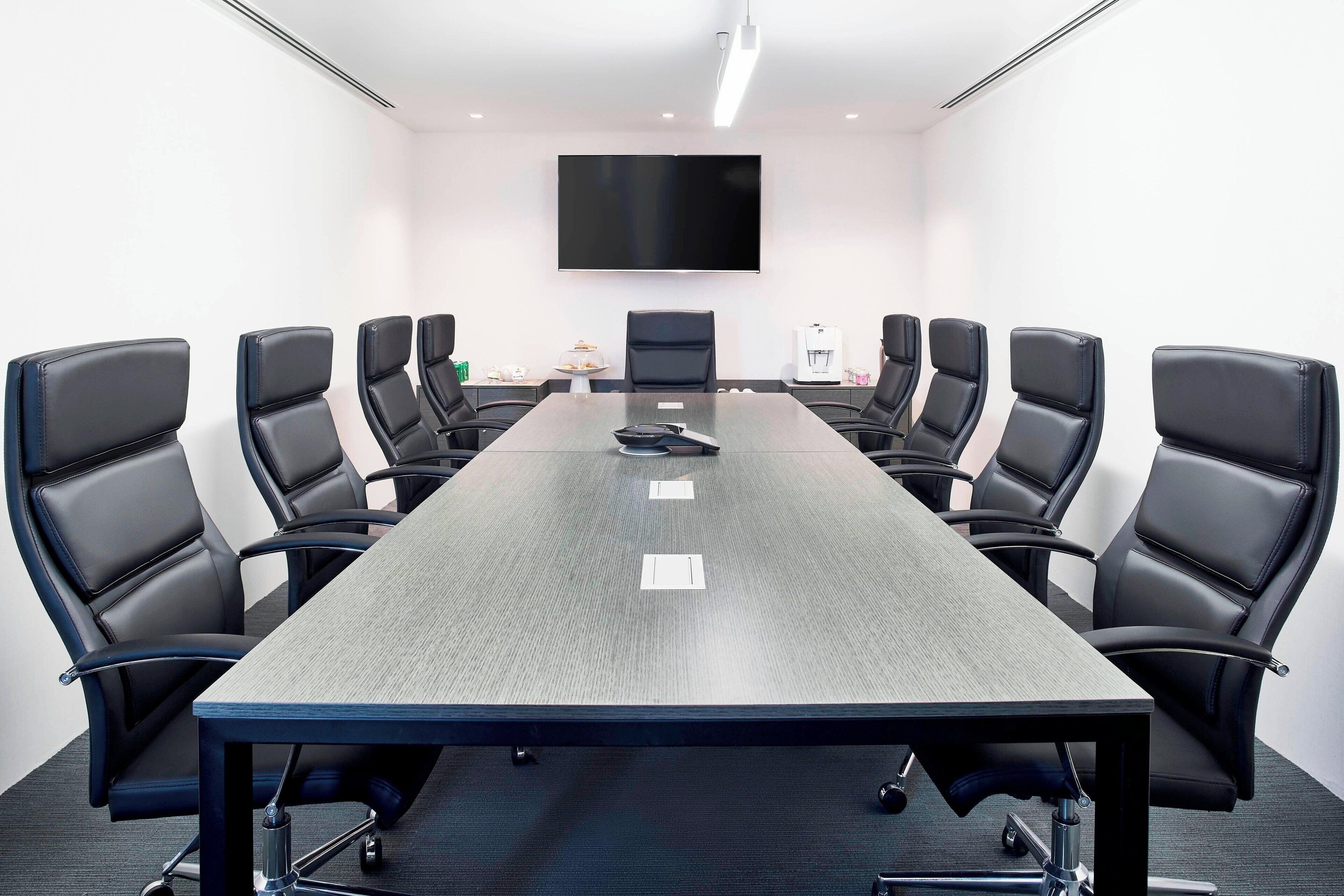 A conference room with a long table, black chairs, a flat-screen TV, and a coffee machine.