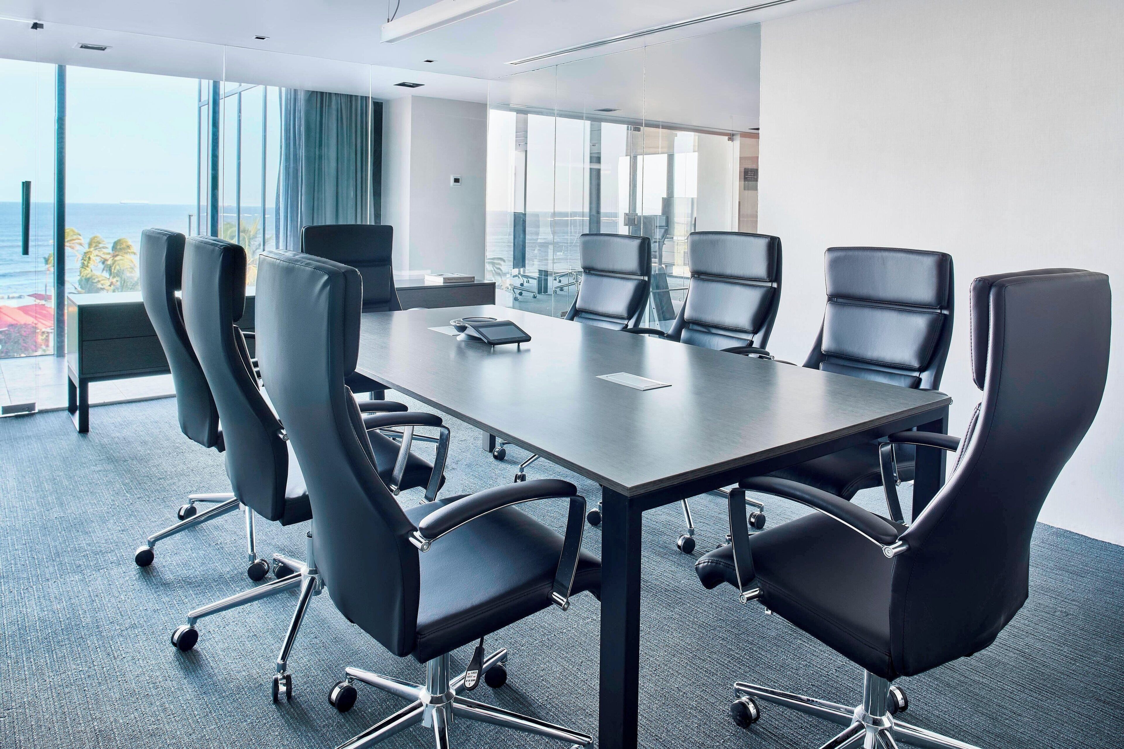 A conference room with a large table and modern chairs, large windows with a view of the ocean, and a glass partition.