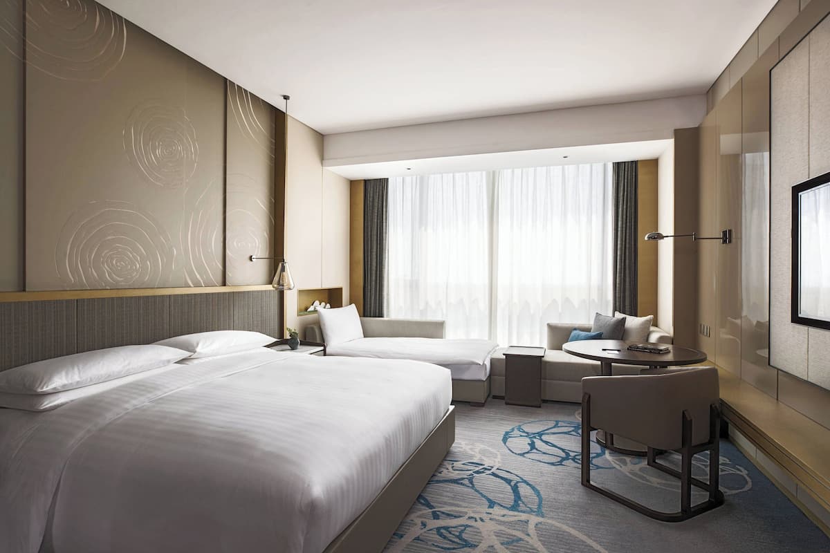 Une chambre d’hôtel avec deux lits, un petit coin salon, une télévision et une grande fenêtre avec des rideaux.