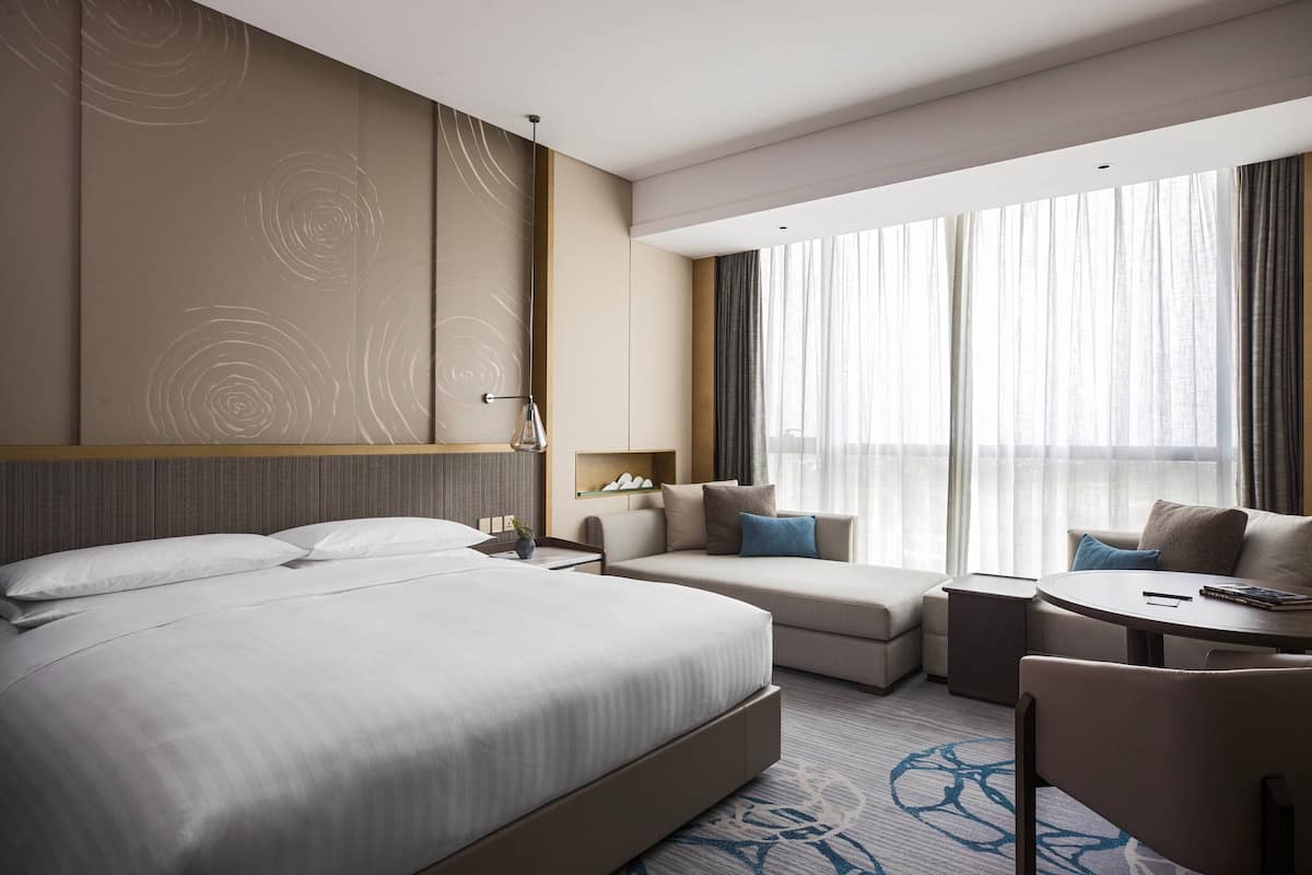 Une chambre d’hôtel moderne dotée d’un grand lit, d’un canapé, d’une petite table et offrant une vue sur la ville grâce à de grandes fenêtres.