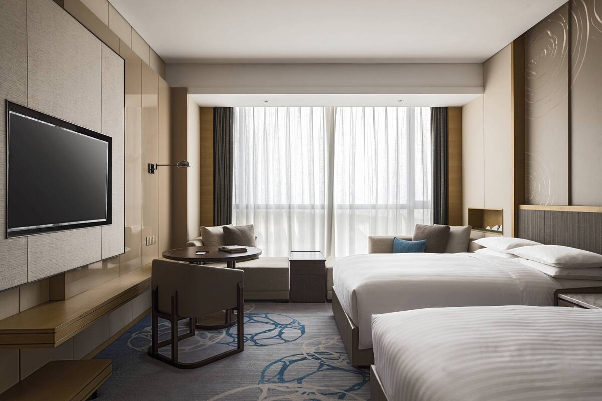 Une chambre d’hôtel dotée d’une grande télévision à écran plat, de deux lits, d’un coin salon avec une petite table et d’une grande fenêtre avec des rideaux légers.