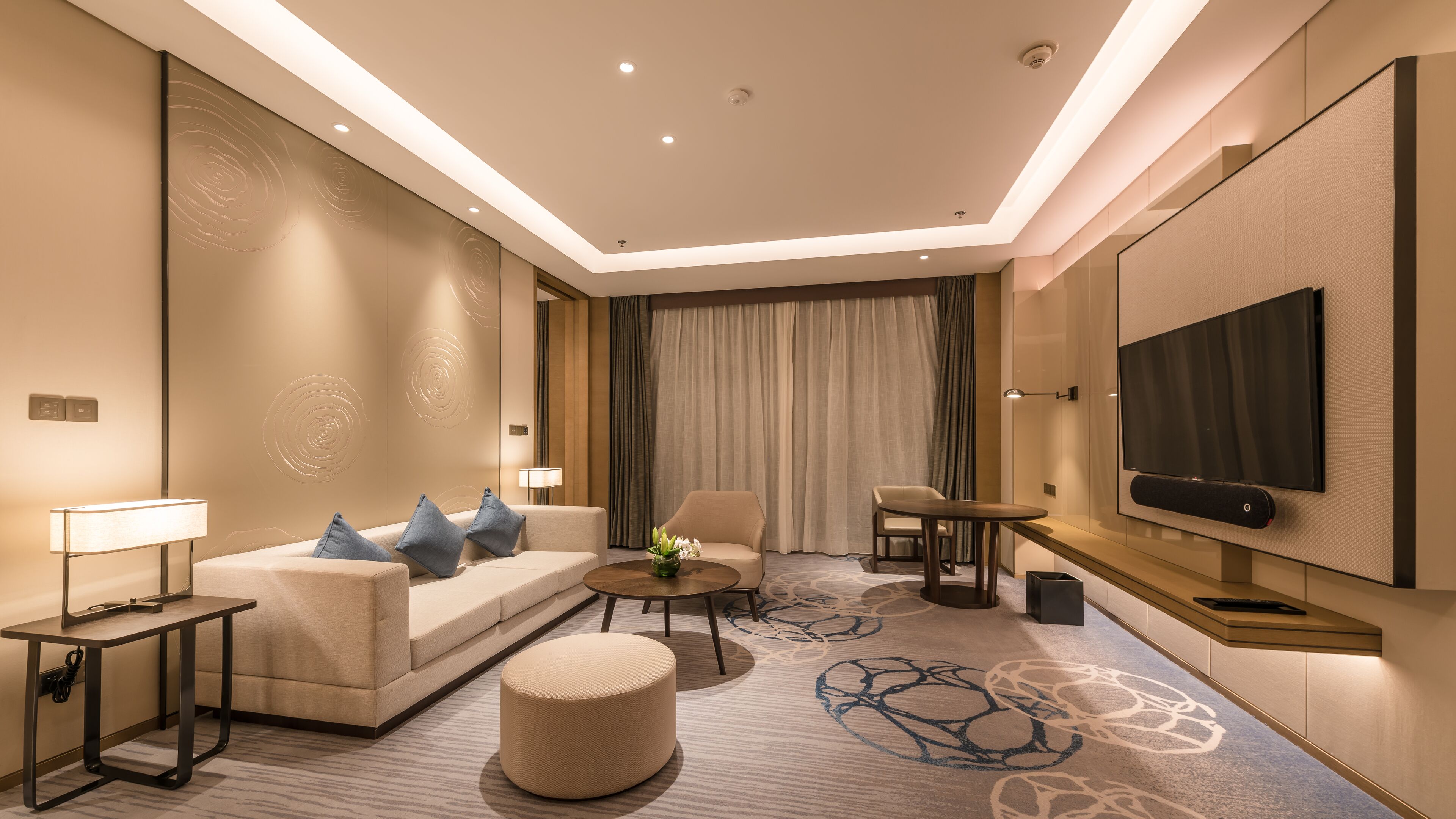 A modern hotel room with a sofa, armchair, ottoman, coffee table, and a flat-screen TV mounted on the wall.