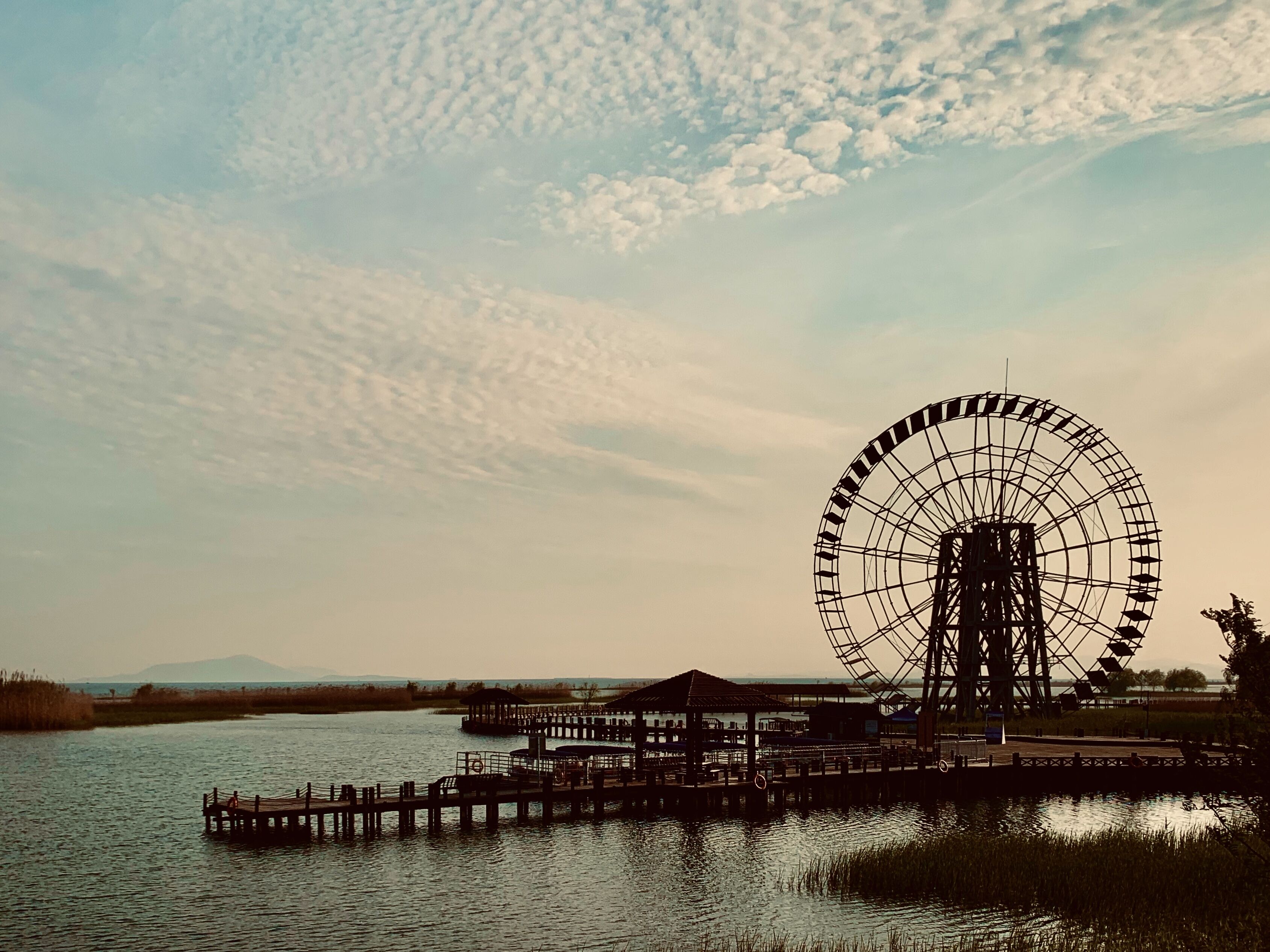 Une grande roue se dresse près d’une étendue d’eau, avec une jetée et des bâtiments en arrière-plan.