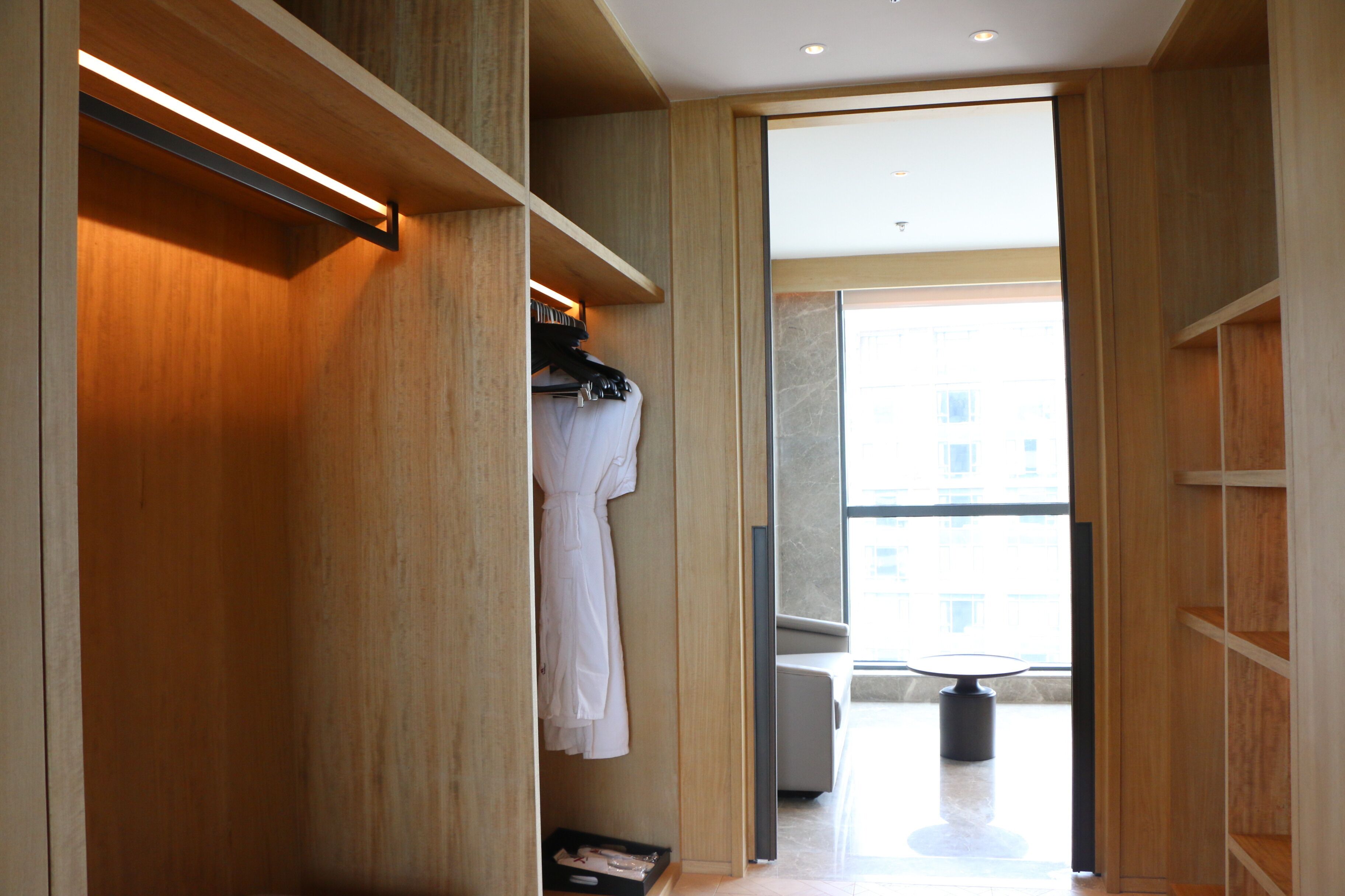 Een moderne hotelkamer met een houten garderobe, een zithoek met een tafel en stoelen, en een groot raam met uitzicht op de stad.