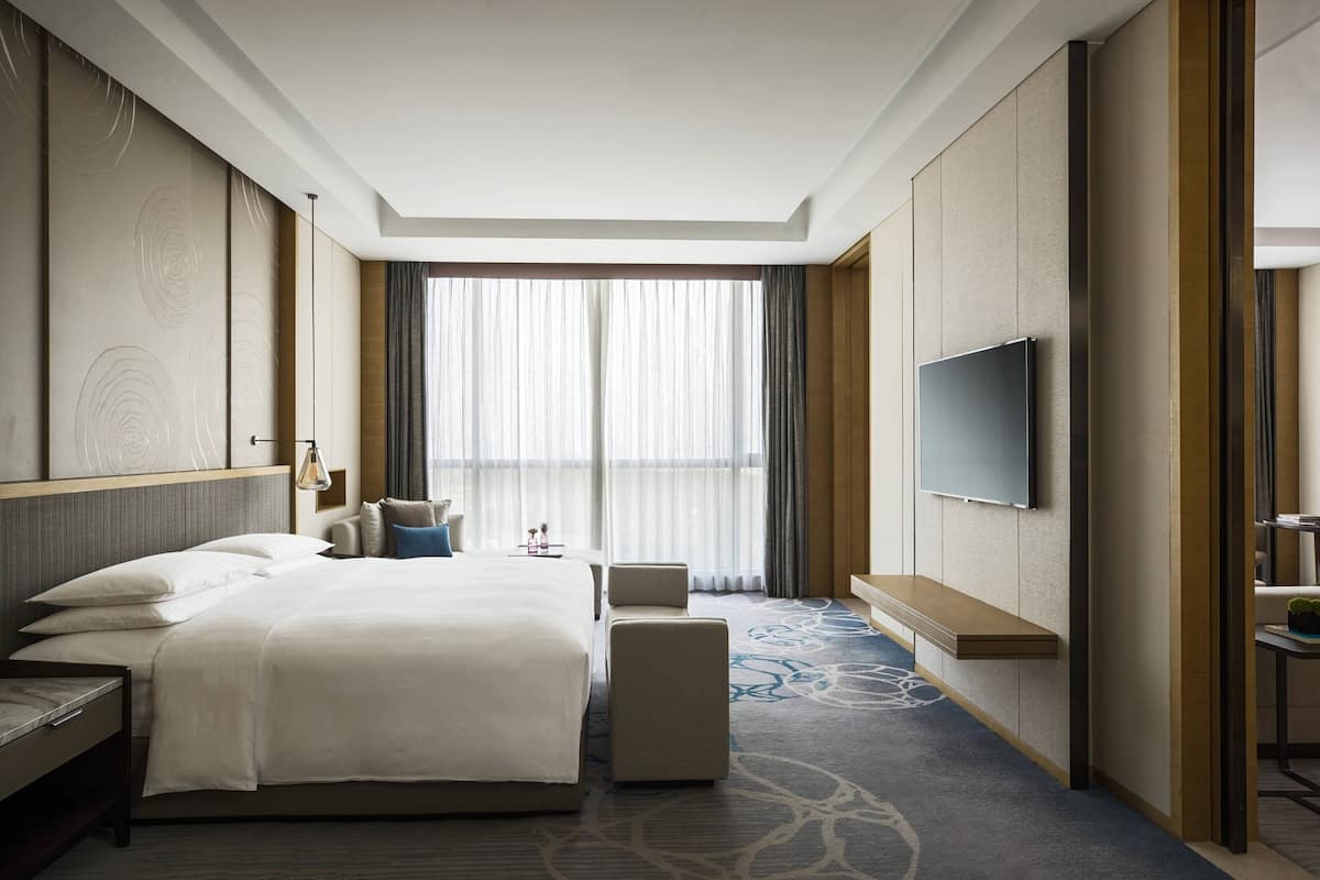 Une chambre d’hôtel dotée d’un grand lit, d’un téléviseur fixé au mur, d’un coin salon avec un banc et offrant une vue sur l’extérieur à travers des rideaux légers.