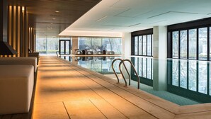 Un espace piscine intérieure moderne, doté d’un long banc, d’une rambarde en verre et de grandes fenêtres offrant une vue sur l’extérieur.