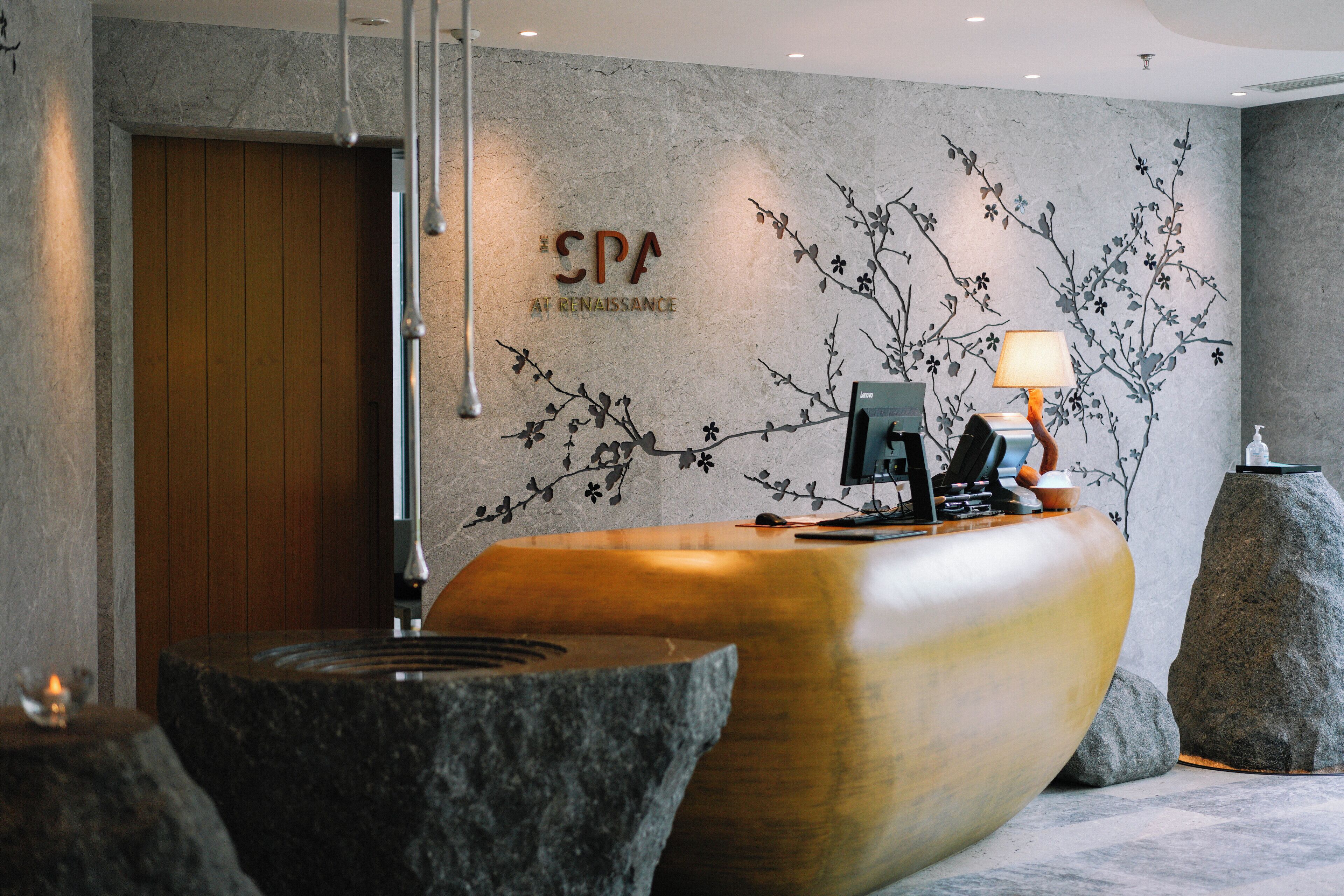 Uma área de recepção com um balcão de madeira, uma placa na parede com a inscrição “SPA AT RENAISSANCE” e uma luminária moderna.
