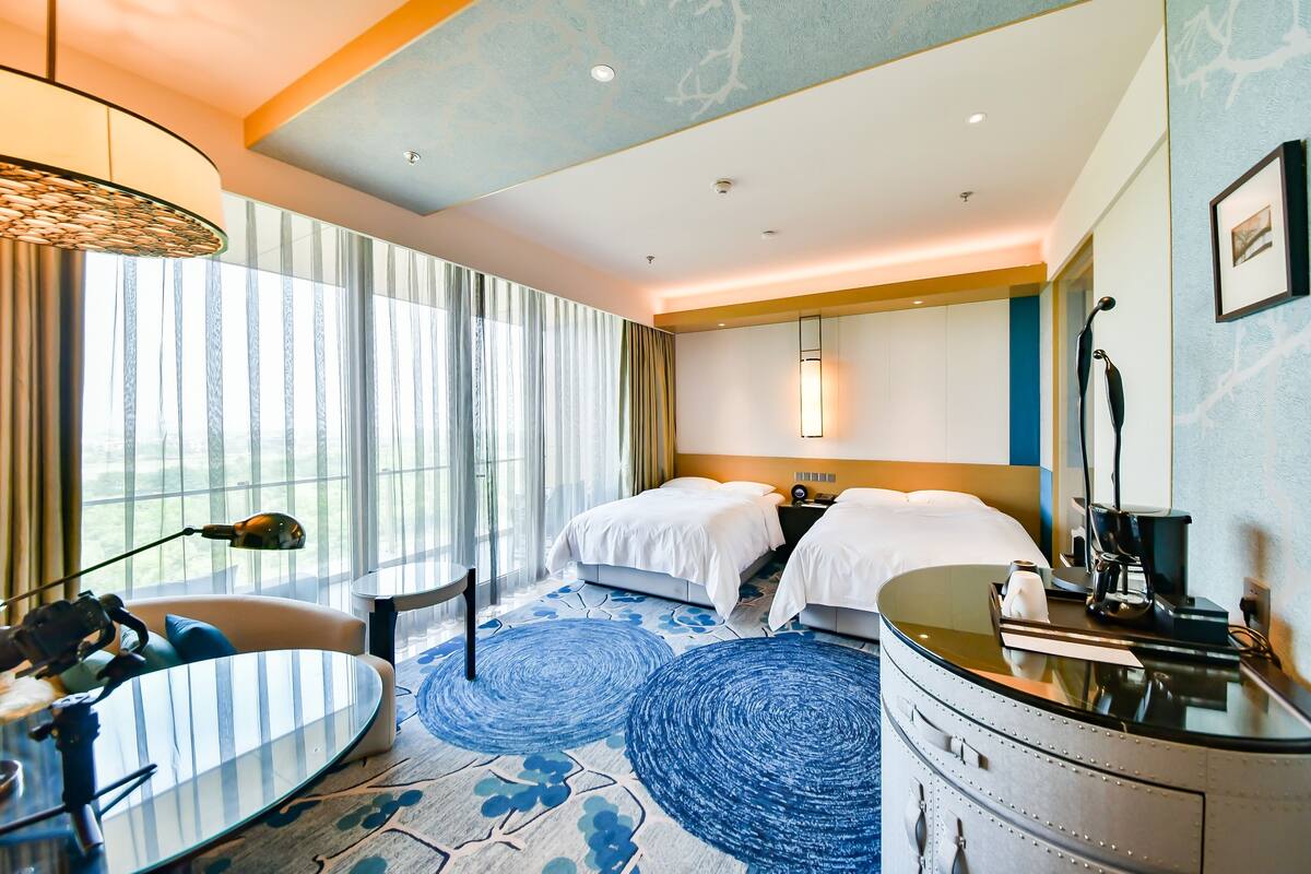 A hotel room with two beds, a desk with a chair, a round table, a large window with curtains, a ceiling light, and a wall-mounted lamp.