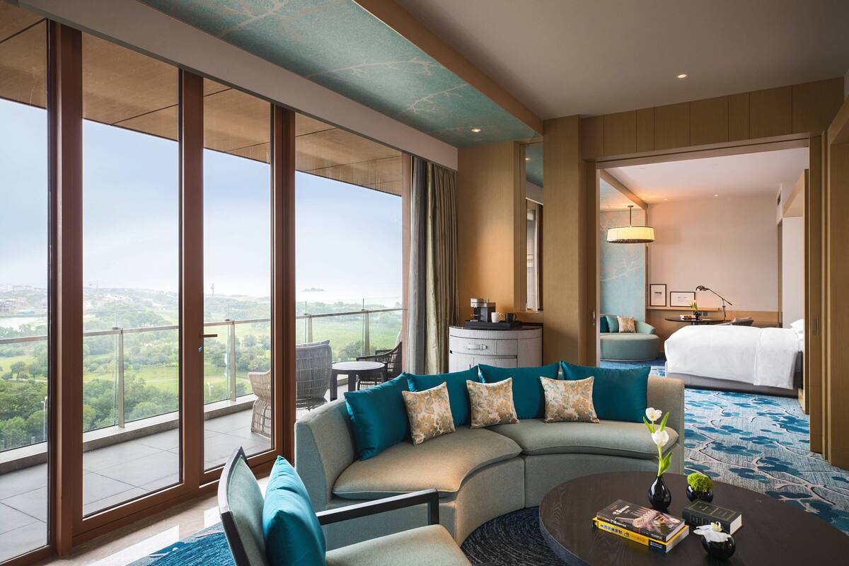 A modern hotel room with a curved sofa, a coffee table with books, a view of the outdoors, a bed, and a bathroom visible through an open door.
