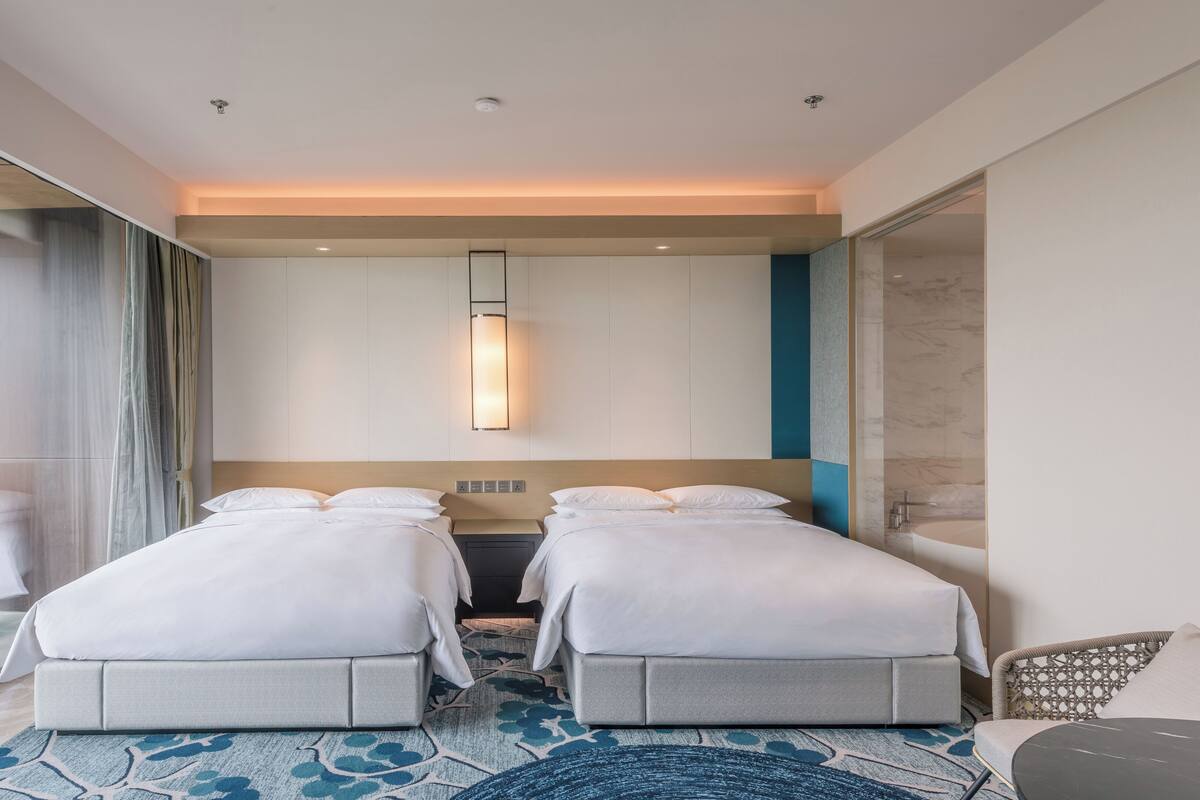A modern hotel room with two beds, a large window with curtains, a wall-mounted light, and a view of the outdoors.