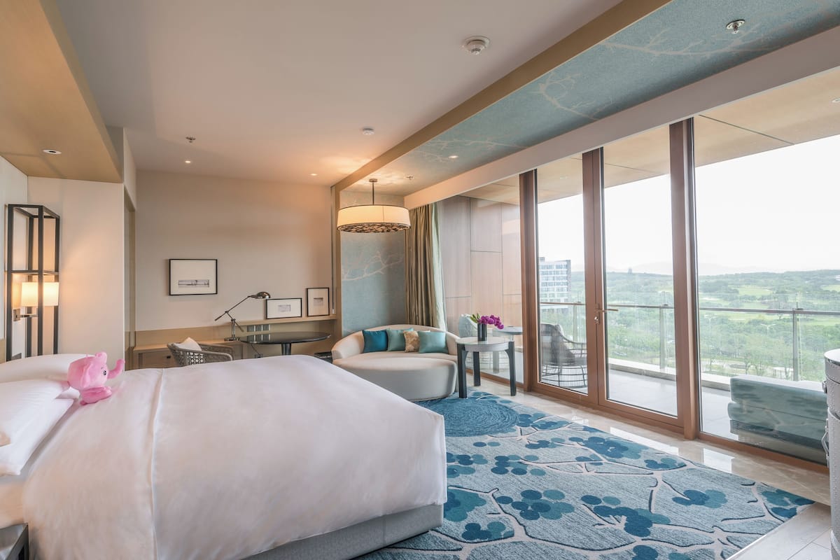 A modern hotel room with a large bed, a desk, a chair, and a balcony with a city view.