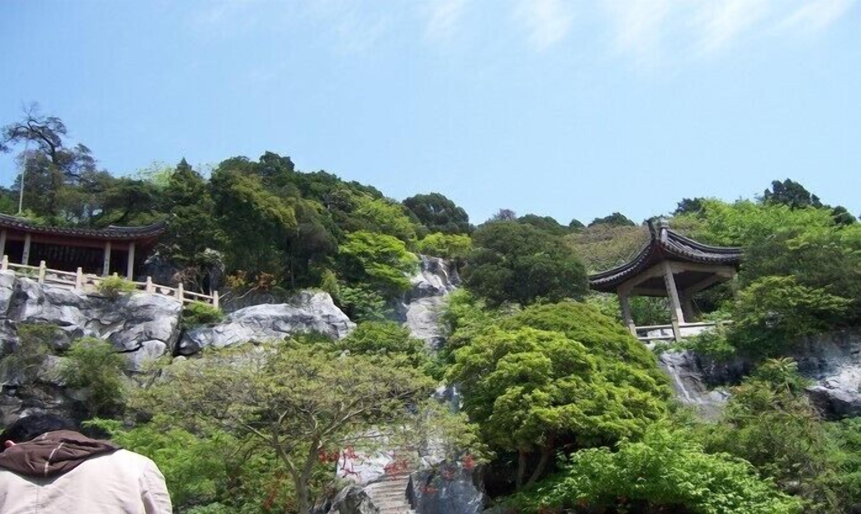 A traditional Chinese pavilion on a rocky hillside with lush greenery and a clear sky.