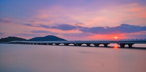 Eine lange Brücke überspannt ruhiges Wasser, im Hintergrund ein Sonnenuntergang.