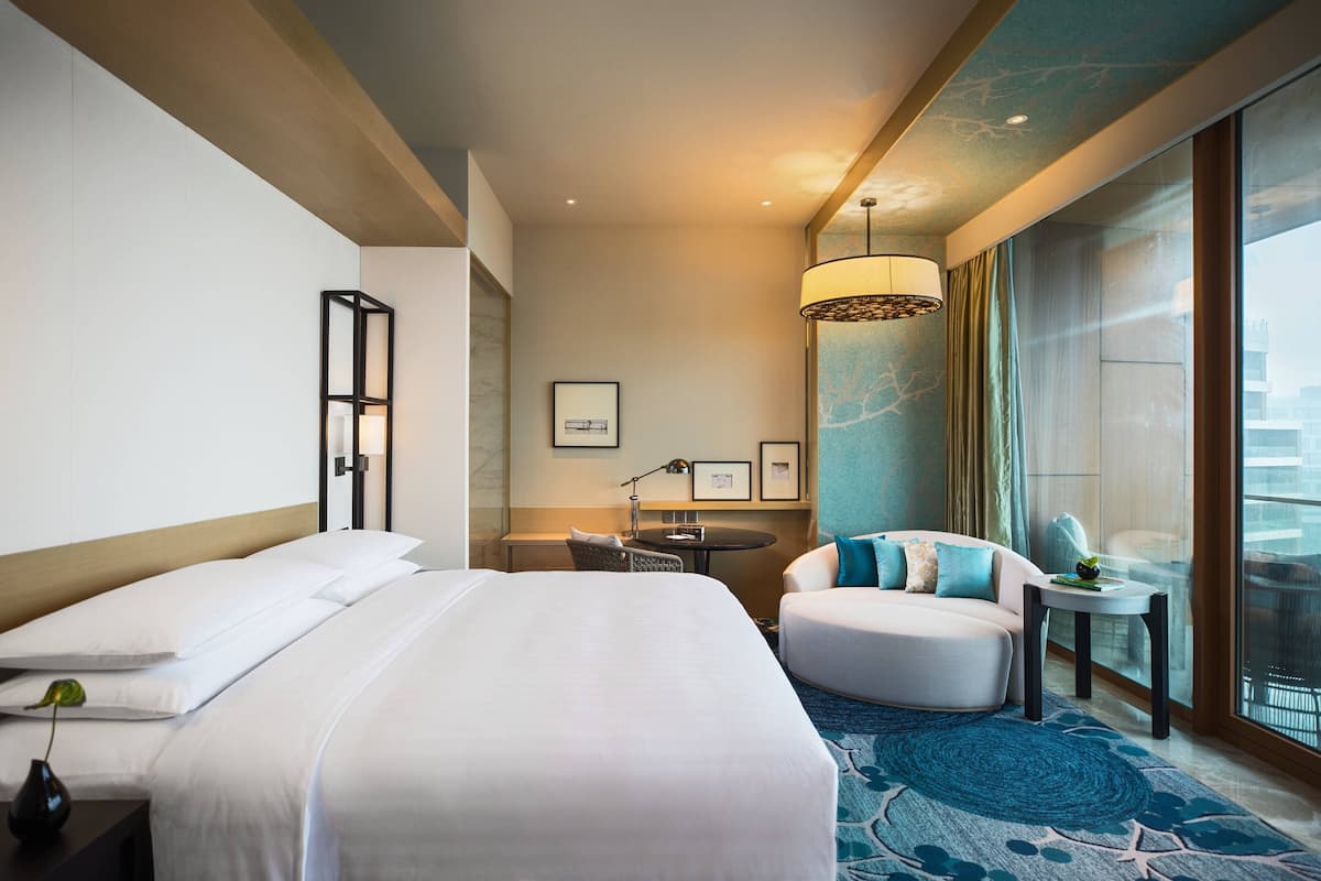 A hotel room with a large bed, a seating area with a sofa and armchairs, a small table, and a view of the city through large windows.