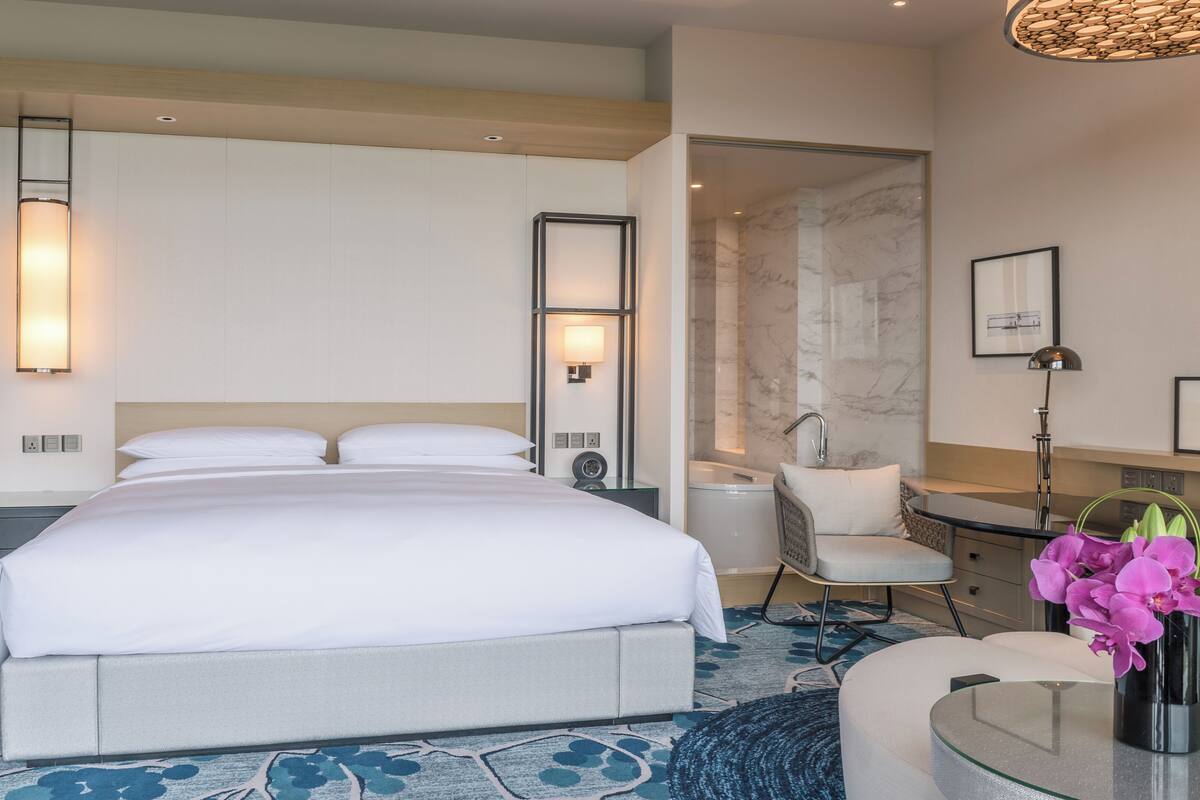 A modern hotel room with a large bed, a sitting area with a chair, a small table, and a bathroom with a marble wall and sink.