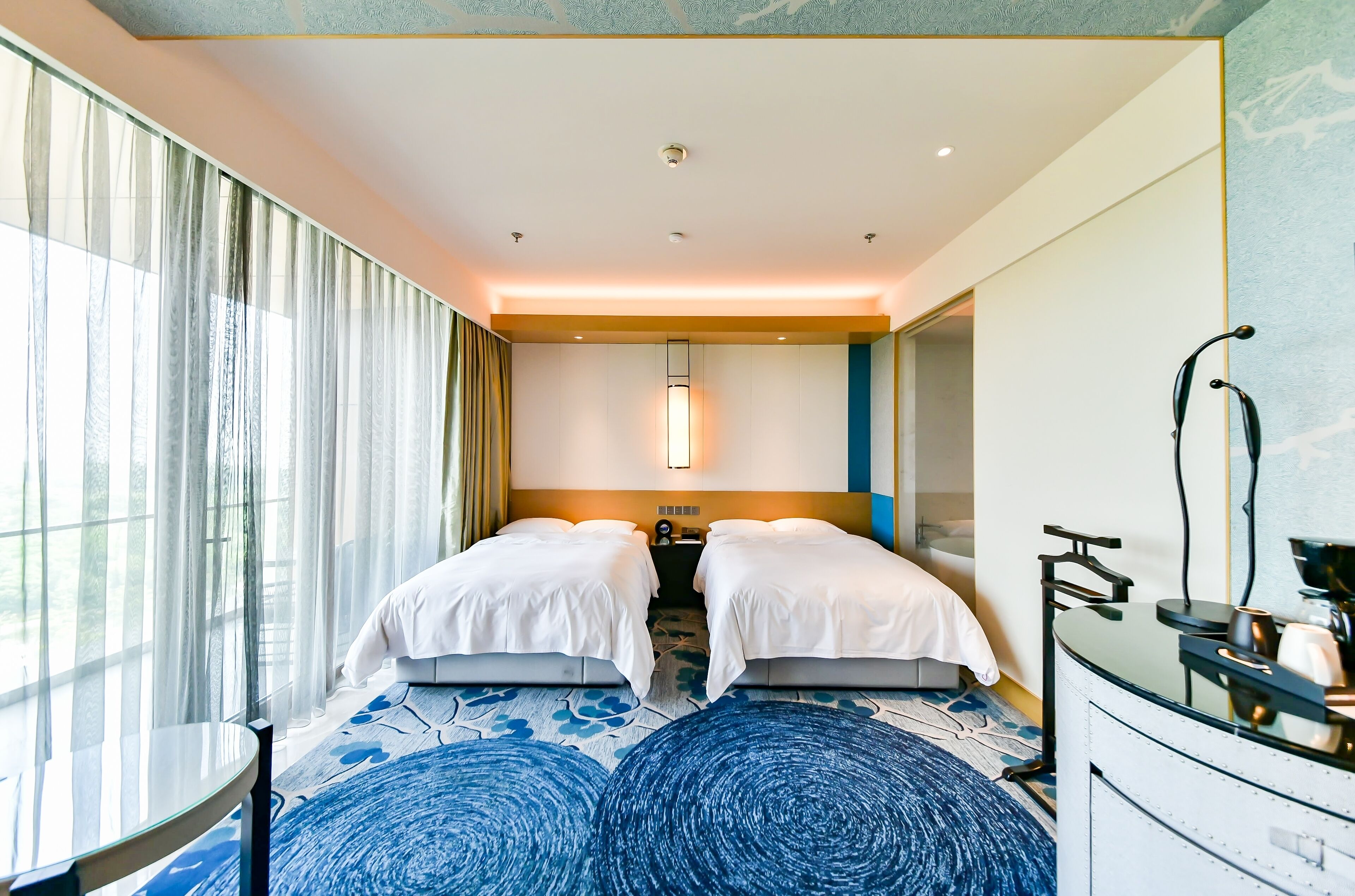 Habitación de hotel con dos camas, una mesita redonda, minibar y un amplio ventanal con cortinas.