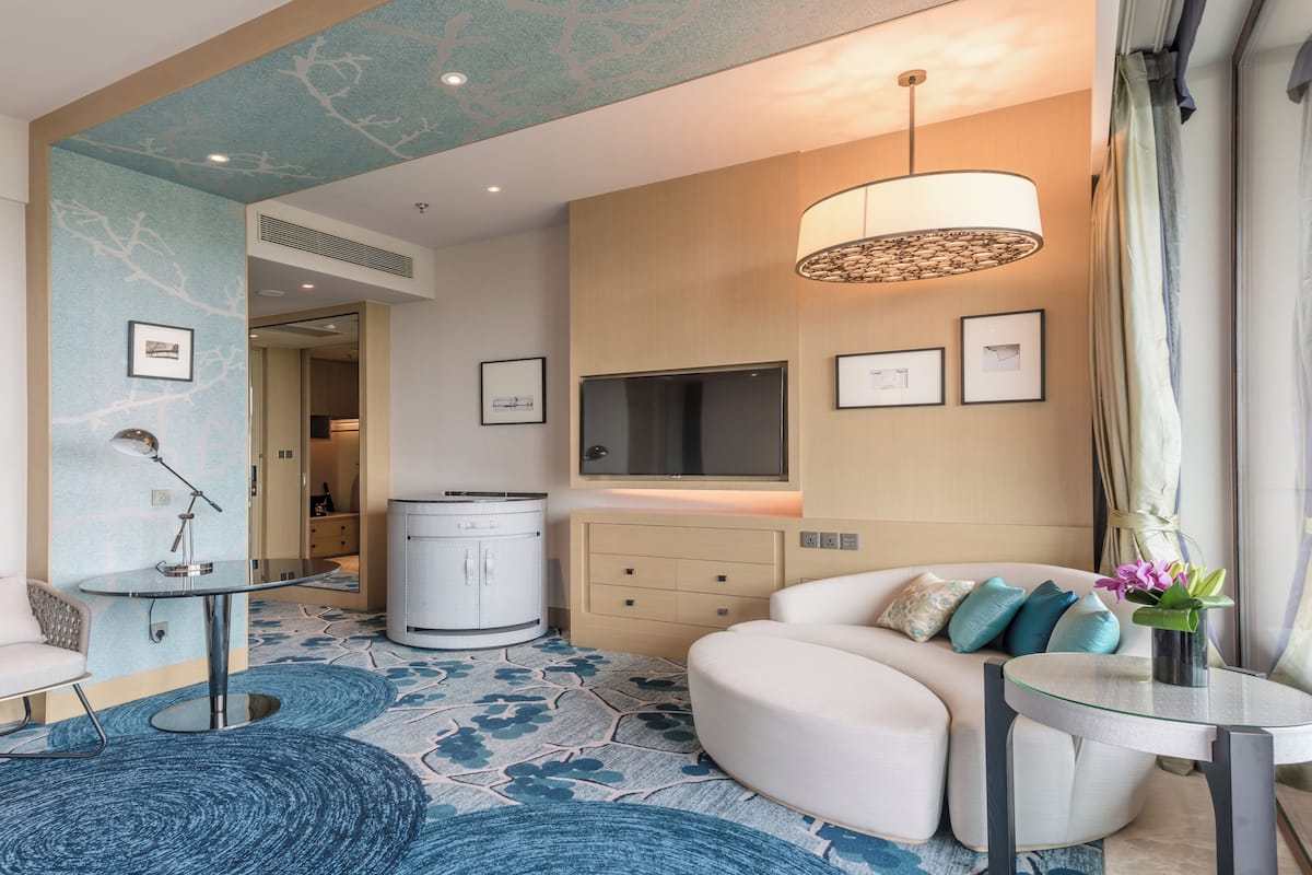 A modern hotel room with a round coffee table, a sofa, a TV mounted on the wall, and a large window with curtains.