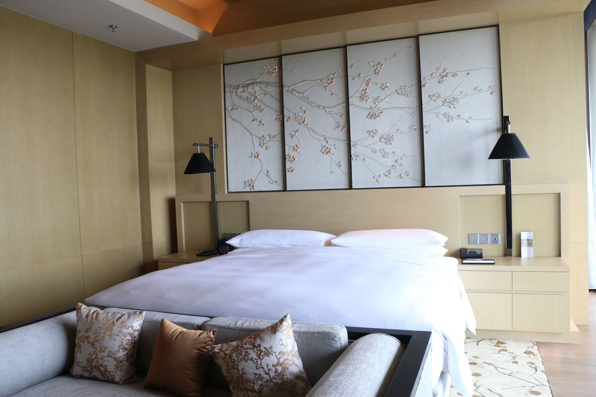 A modern hotel room with a large bed, a sofa with decorative pillows, a bedside table with a lamp, and a wall-mounted artwork.