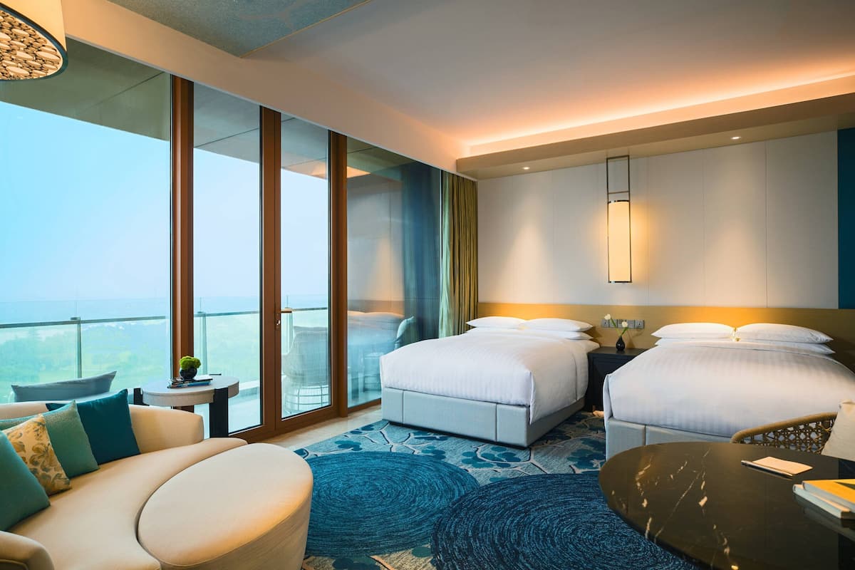 A hotel room with two beds, a sofa, a small table, and a balcony with a view.