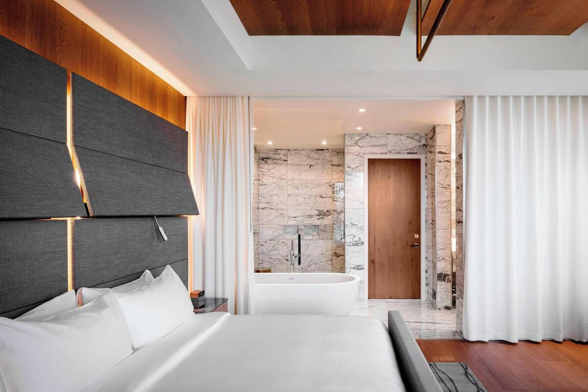 A modern bedroom with a large bed, a built-in headboard, a bathroom with a marble wall, and a wooden door.