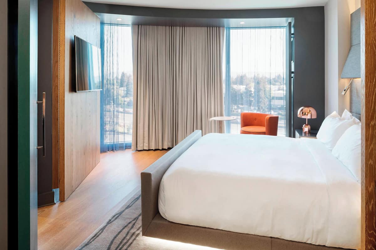 A modern hotel room with a large bed, a small table, a chair, and a TV mounted on the wall.