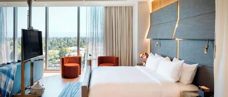 Une chambre d’hôtel moderne dotée d’un grand lit, d’un téléviseur fixé sur un meuble, d’un coin salon avec un fauteuil et une petite table, et offrant une vue sur l’extérieur grâce à de grandes fenêtres.