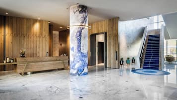 A modern lobby with a marble floor, a wooden reception desk, a large pillar with a geometric design, and a staircase with a blue carpet.