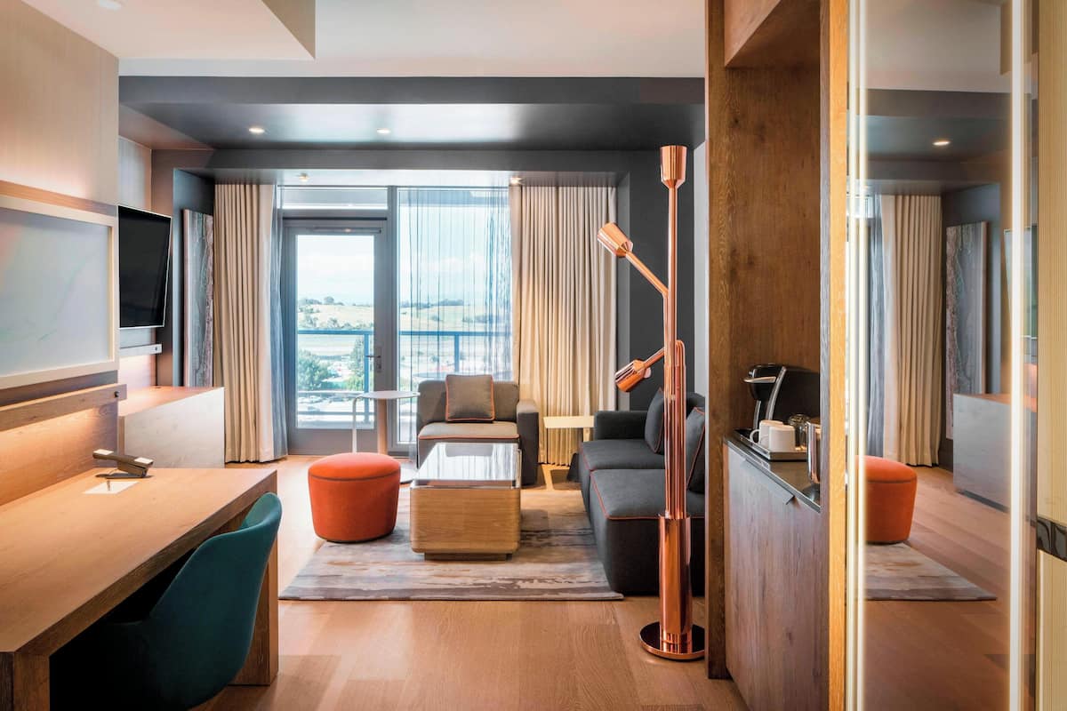 A modern hotel room with a wooden desk, a sofa, a large window with curtains, and a view of the city.