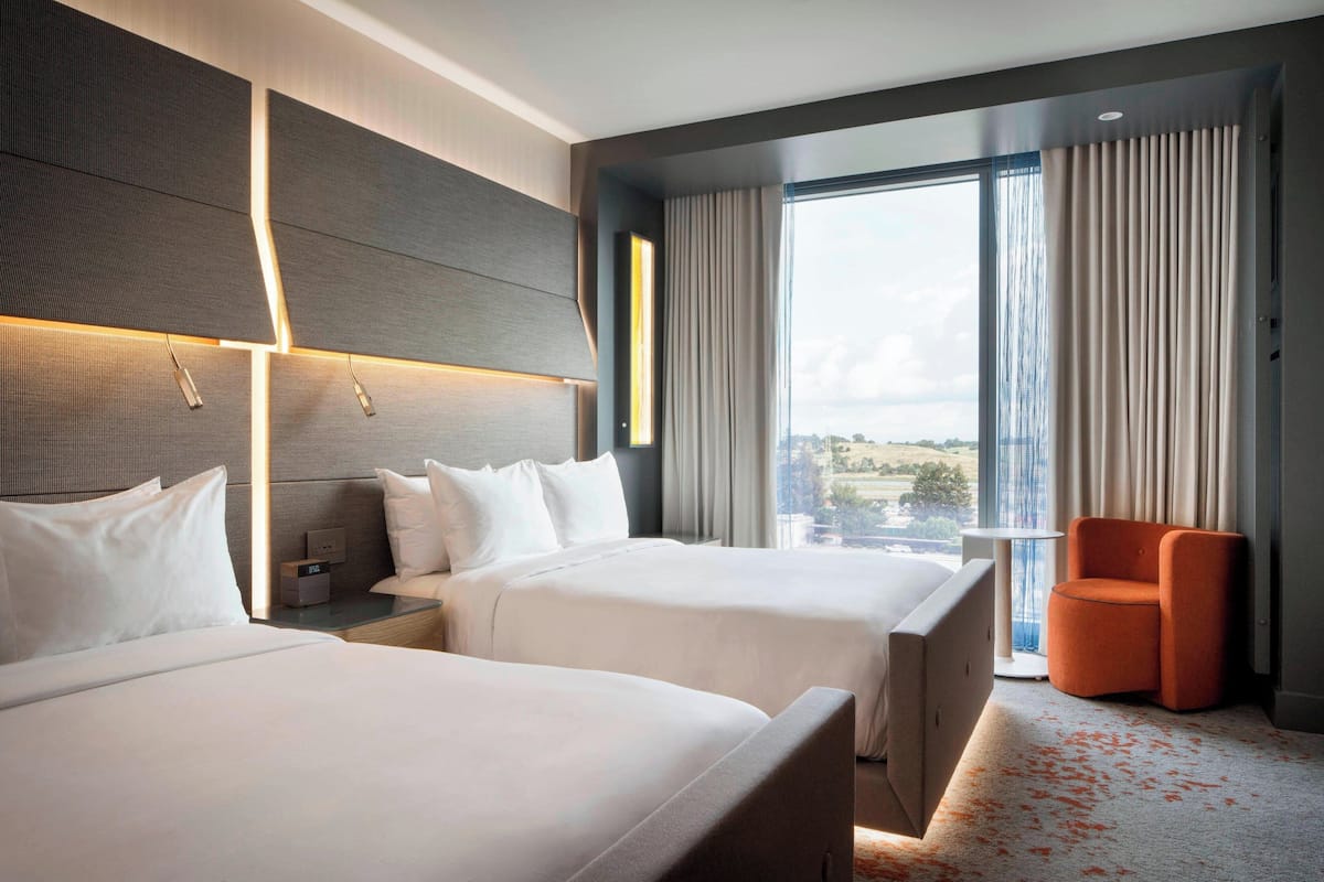 A hotel room with two beds, a small table, a chair, and a large window with curtains.