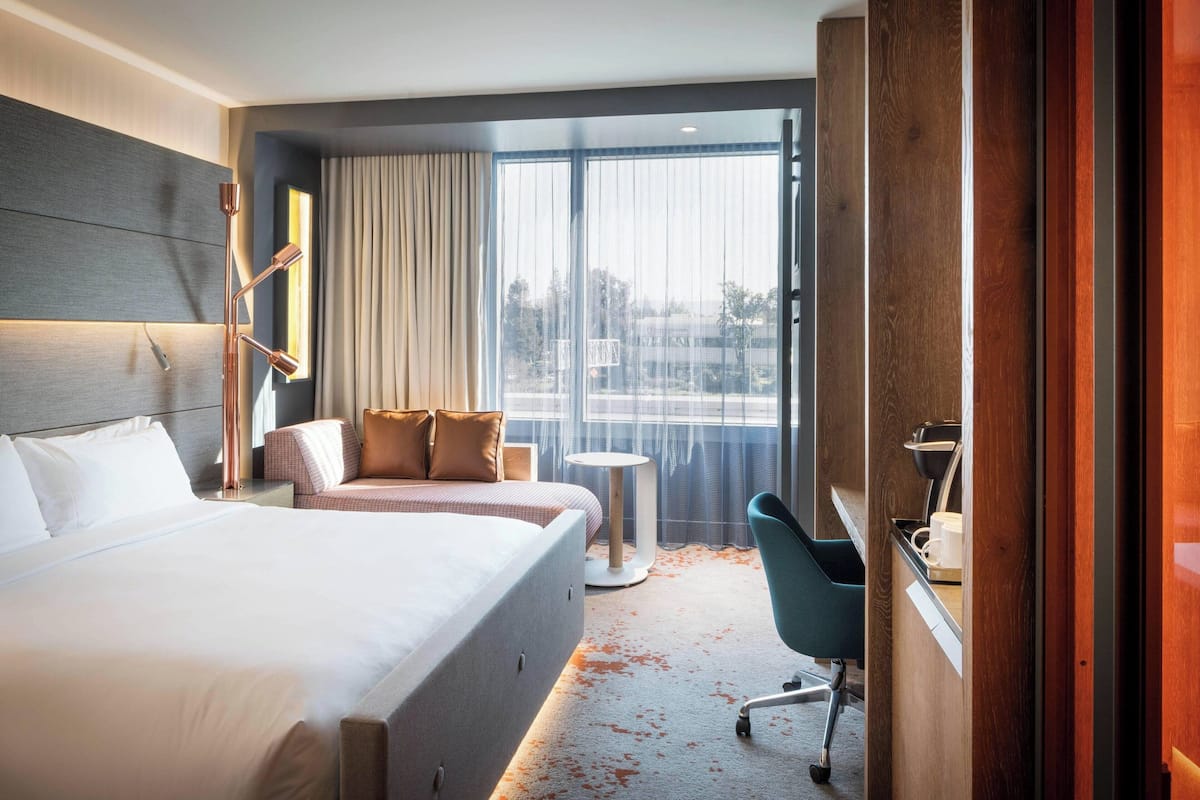 A modern hotel room with a large bed, a desk, a chair, and a view of the city through large windows.