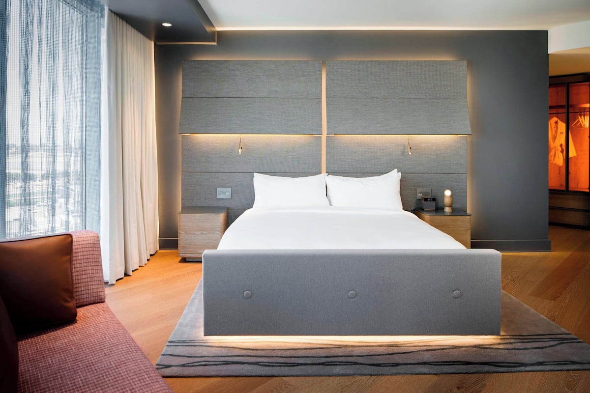 A modern hotel room with a large bed, a nightstand, a lamp, and a view of the city through the window.