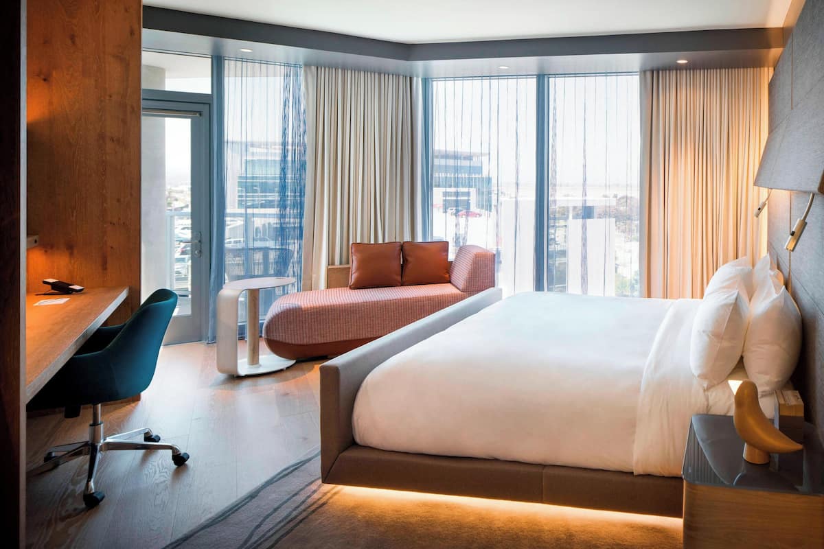 A modern hotel room with a large bed, a desk with a chair, a small sofa, and large windows offering a city view.