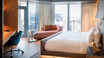 A modern hotel room with a large bed, a desk with a chair, a small sofa, and large windows offering a city view.
