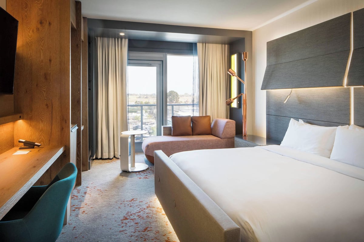 A hotel room with a large bed, a desk, a chair, a small table, and a sofa. There is a window with curtains and a view of the city.