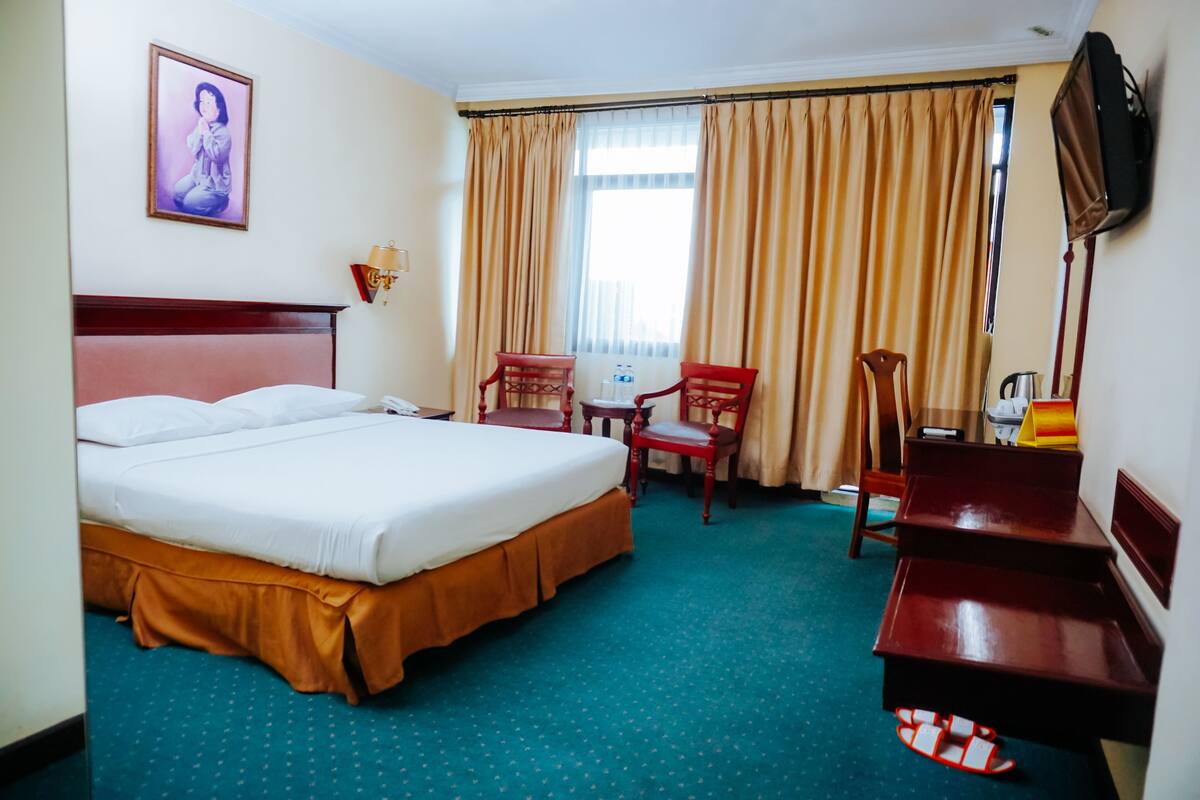 Ein Hotelzimmer mit einem großen Bett, zwei Sesseln, einem Schreibtisch, einem Fernseher und einem Fenster mit Vorhängen.