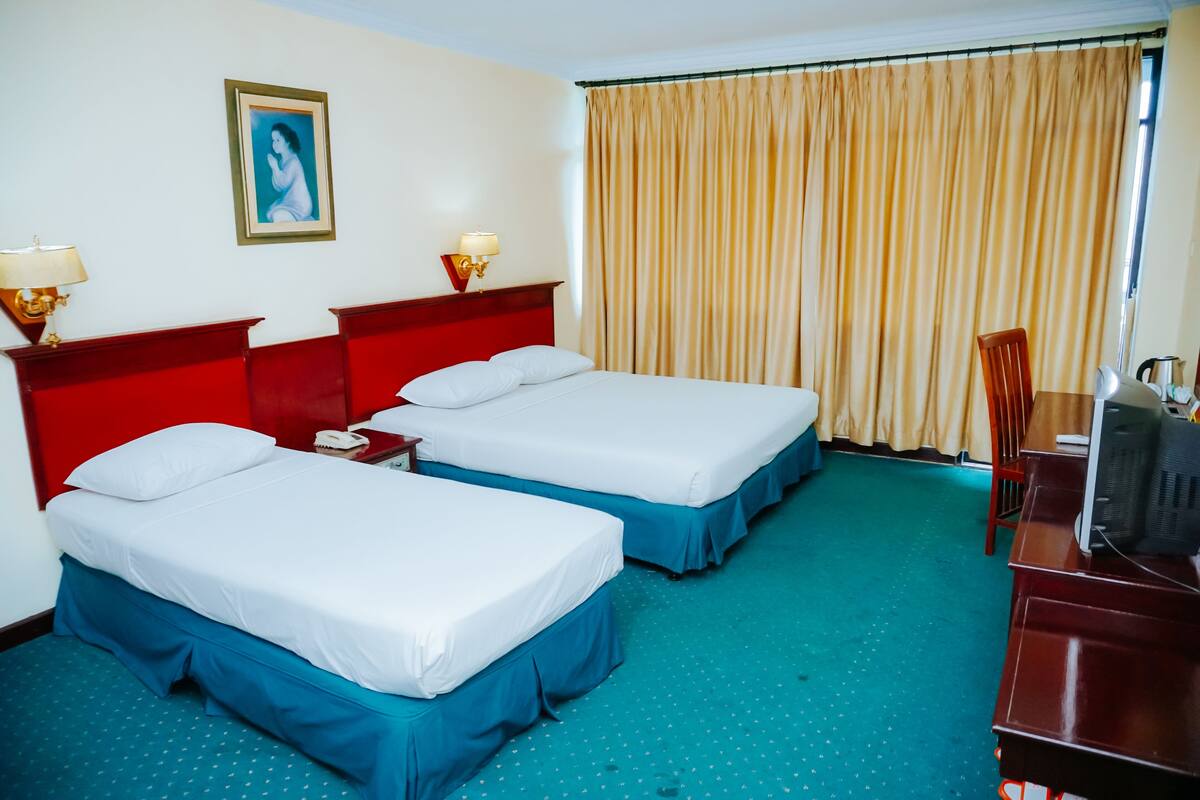 Ein Hotelzimmer mit zwei Betten, einem Schreibtisch, einem Stuhl, einem Fernseher und einem Vorhangfenster.