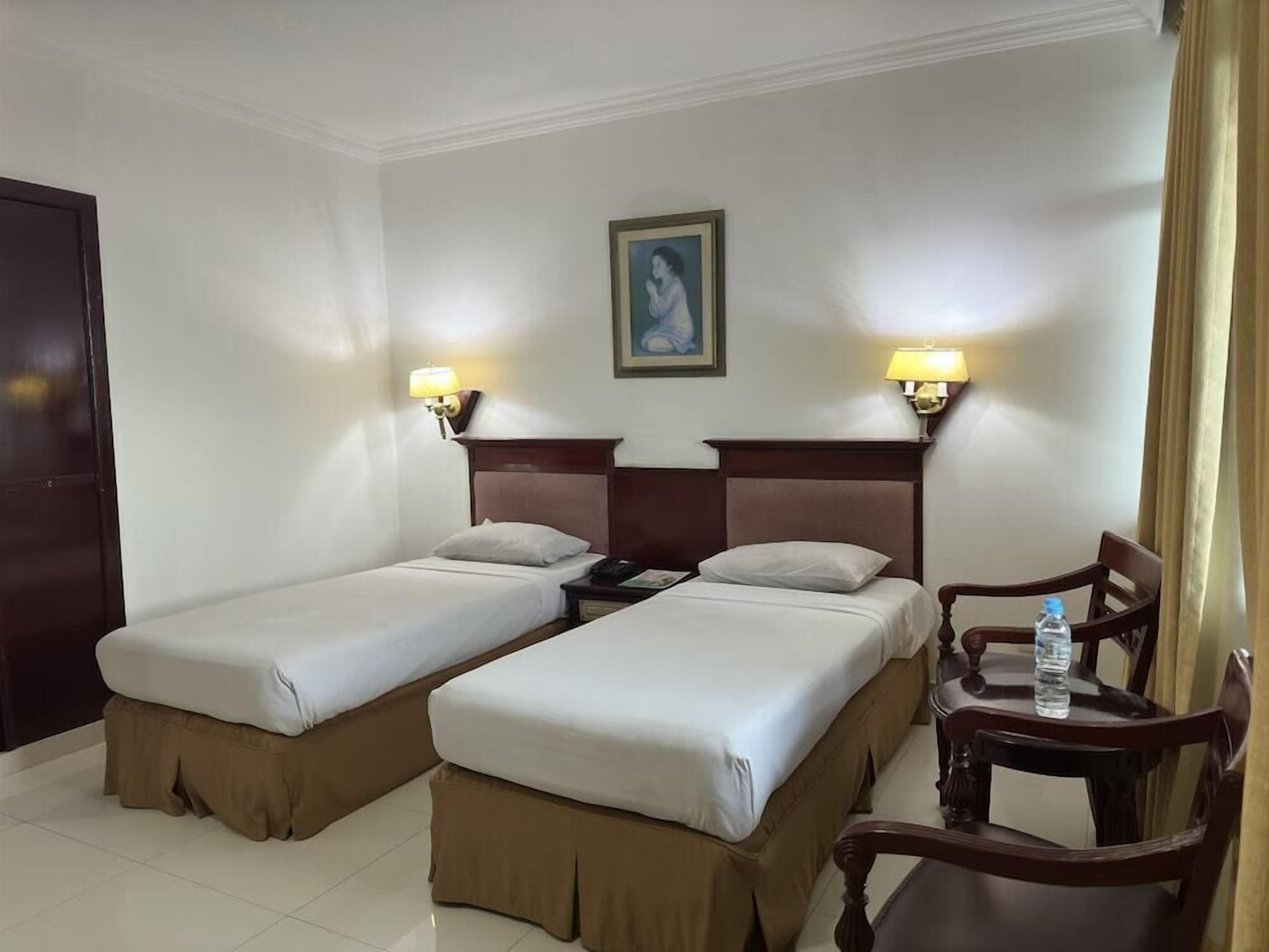A hotel room with two beds, a nightstand with a lamp, a chair, and a table with a water bottle.
