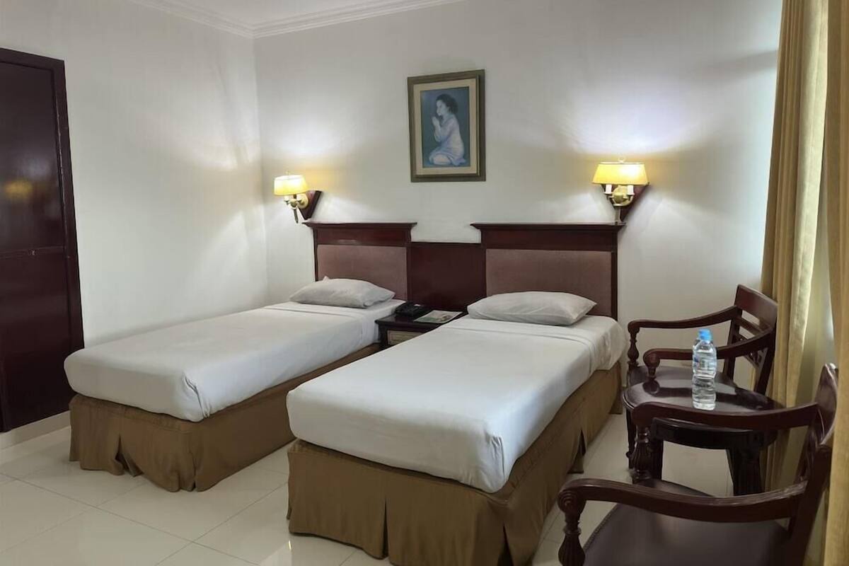 Ein Hotelzimmer mit zwei Betten, einem Nachttisch mit Lampe, einem Sessel und einem Tisch mit einer Wasserflasche.